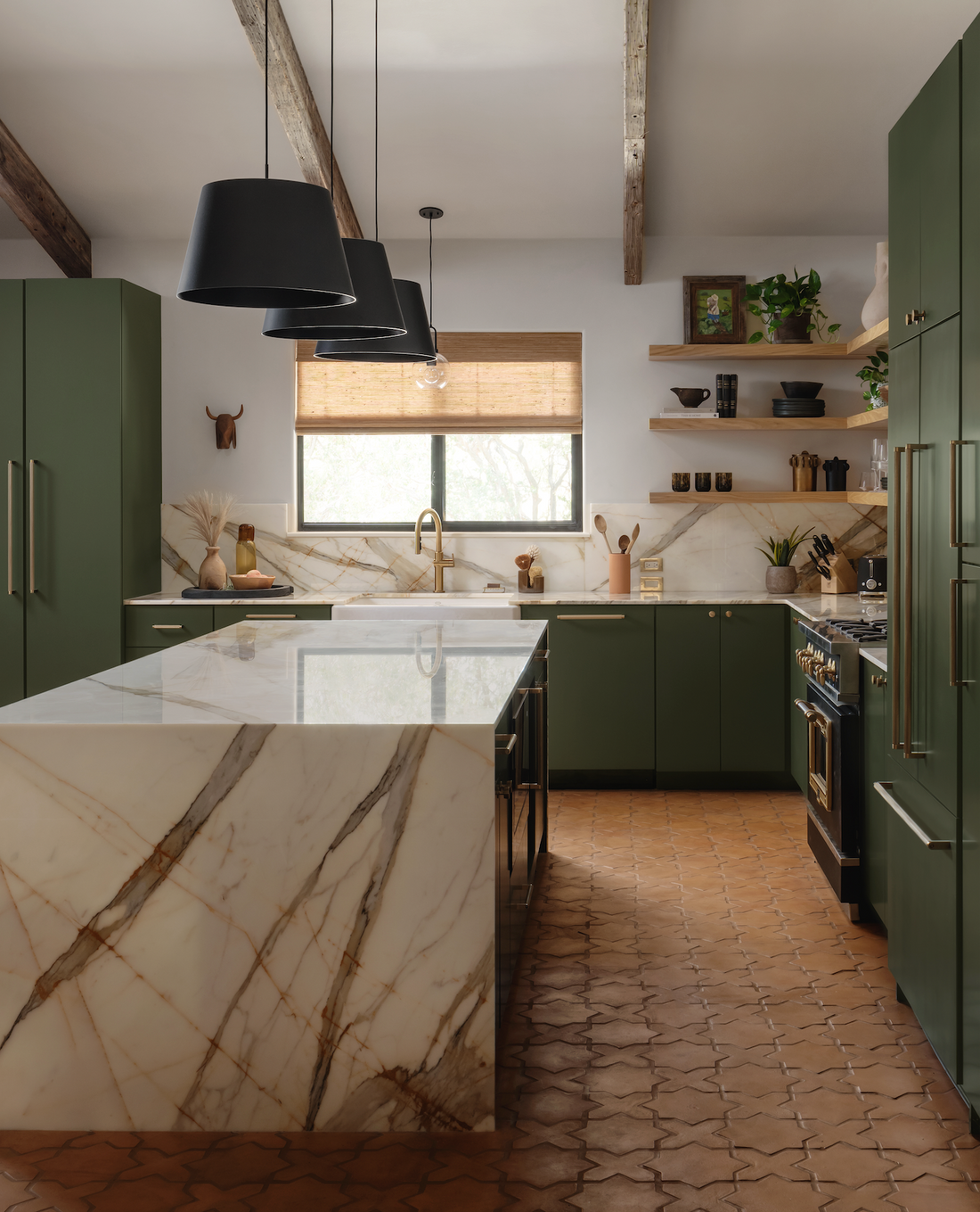 Modern kitchen with green cabinets, large marble island, gold fixtures, open wood shelves with plants and decor, black pendant lights, and Zia Tile Stars & Cross + Madera terracotta flooring. Light streams through a window with a woven shade.