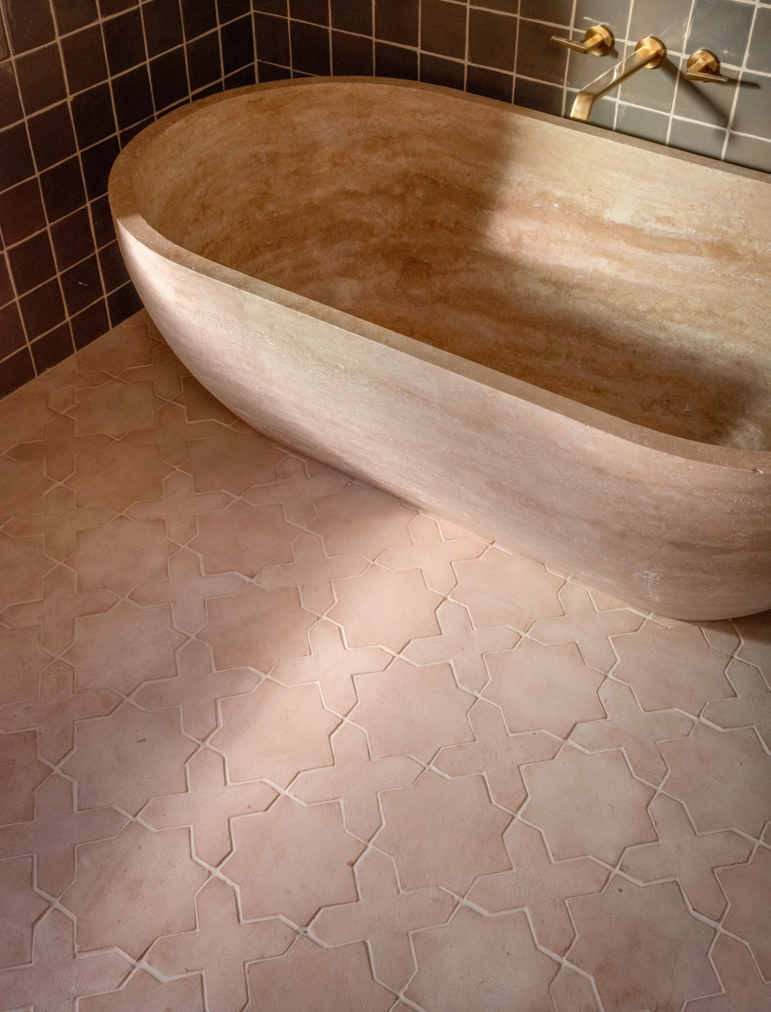A stone bathtub with a natural beige finish sits on Zia Tile’s Stars & Cross + Blanco tiles in a light pink geometric pattern. Dark square wall tiles and brass fixtures accent the space as sunlight softly illuminates the scene.