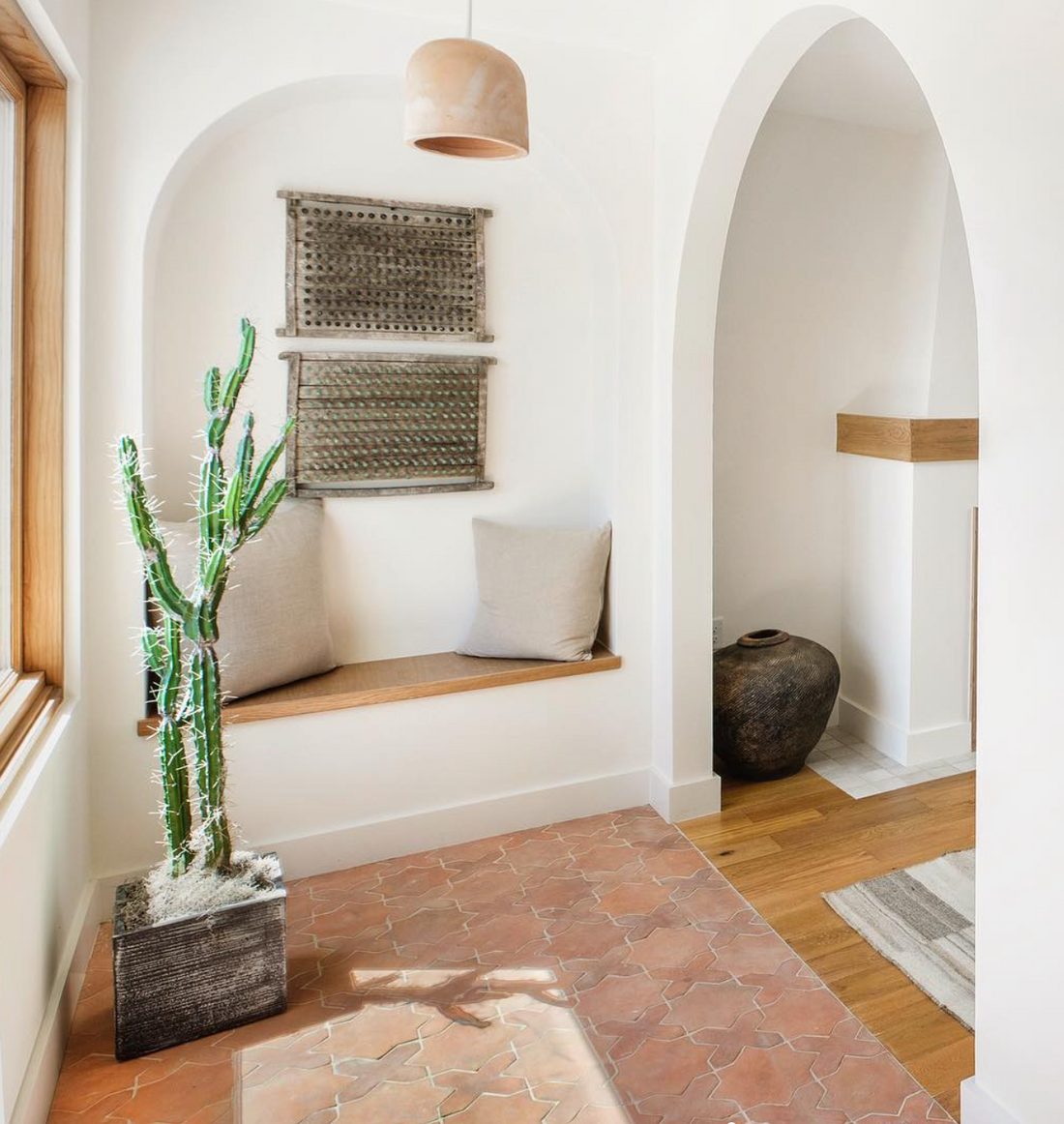 A bright, minimalist entryway featuring Zia Tile’s Stars & Cross + Fired Earth (Just Stars) terracotta tile floor, a tall potted cactus, wooden bench with beige cushions, woven wall art, arched doorway, and a large ceramic vase.