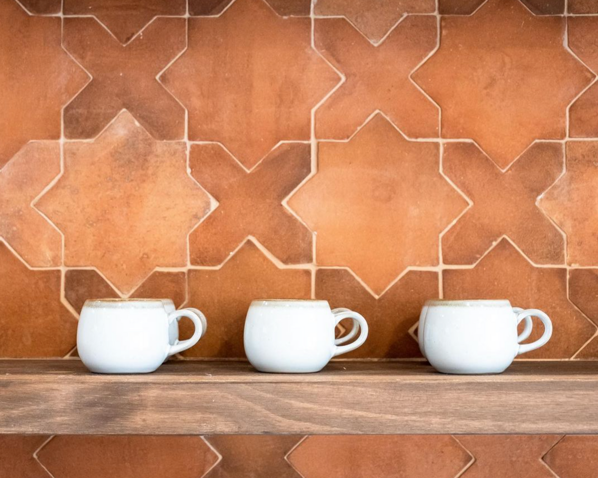 Three white ceramic mugs are lined up on a wooden shelf in front of Zia Tile’s Stars & Cross + Red Clay (Just Stars) handmade geometric tile wall.