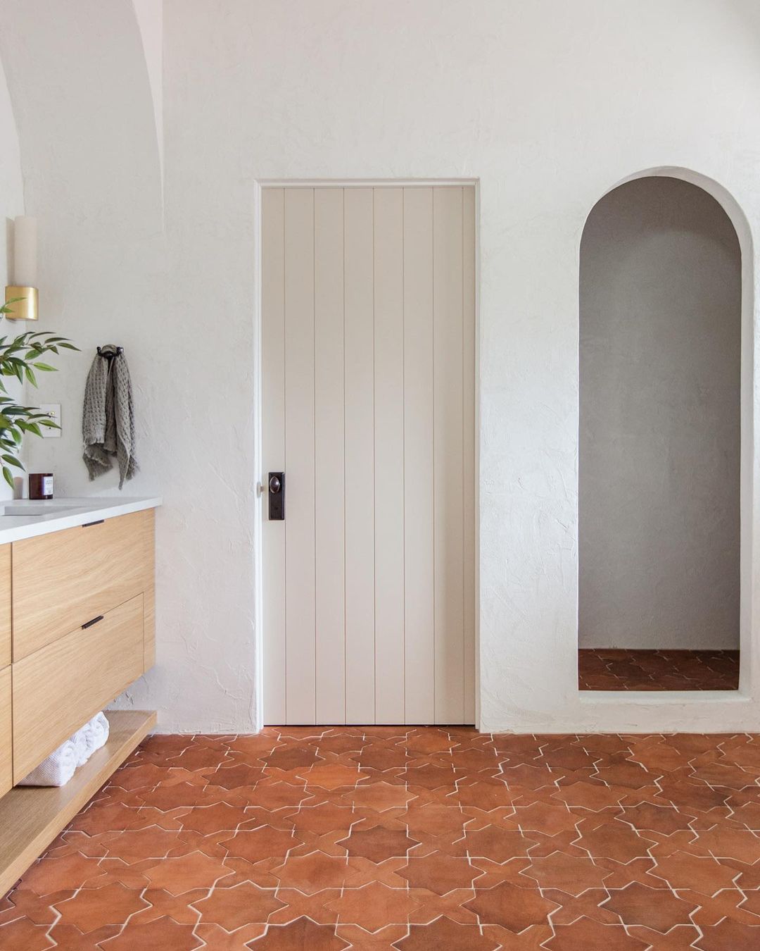 A minimalist bathroom features Zia Tile’s Stars & Cross + Red Clay (Just Stars) terracotta flooring, a light wood vanity with a white countertop, a wall-mounted towel, a beige paneled door, and an arched alcove with matching handmade tile.