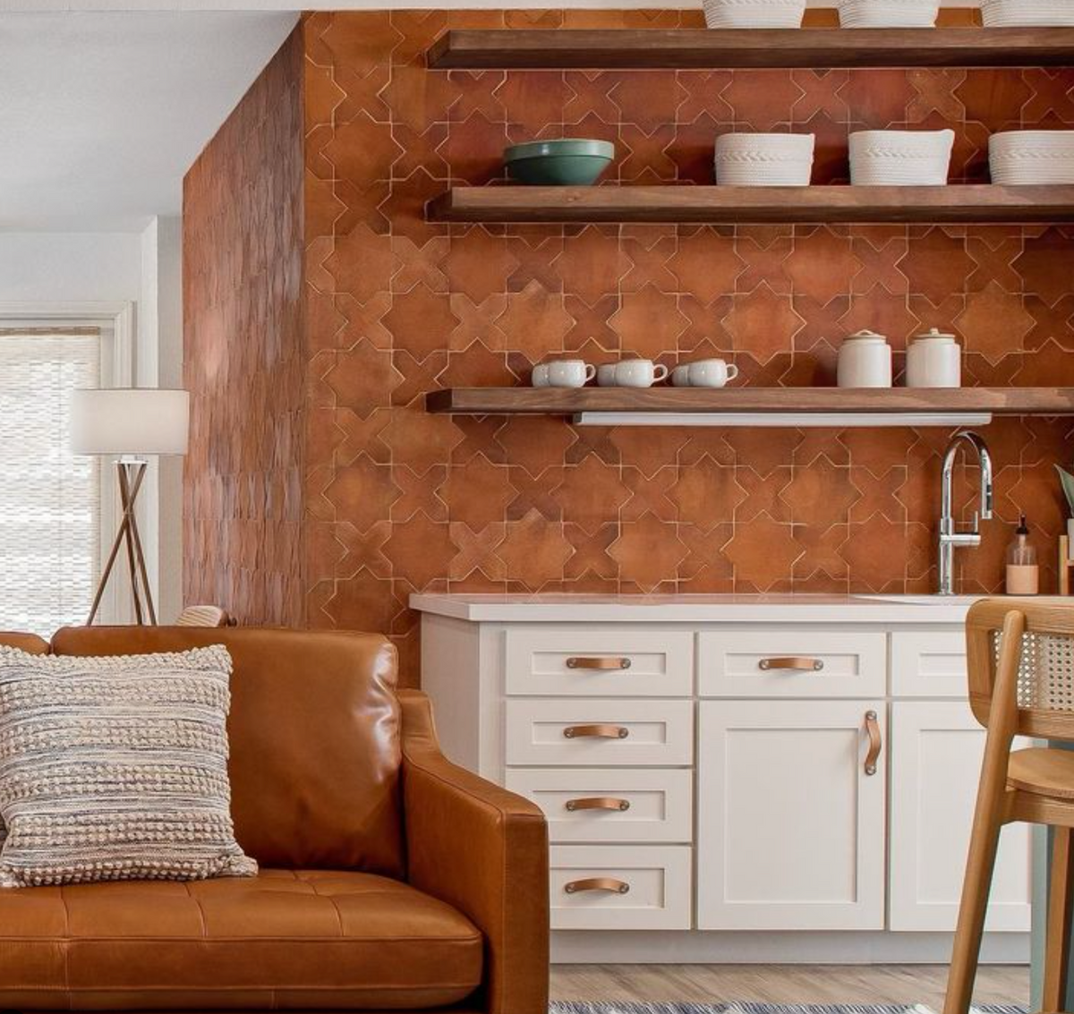 A cozy kitchen features white cabinets, a brown leather sofa, open shelves with white dishes, and a Zia Tile Stars & Cross + Red Clay (Just Stars) cement tile backsplash. A wooden chair and floor lamp add warmth to the inviting space.