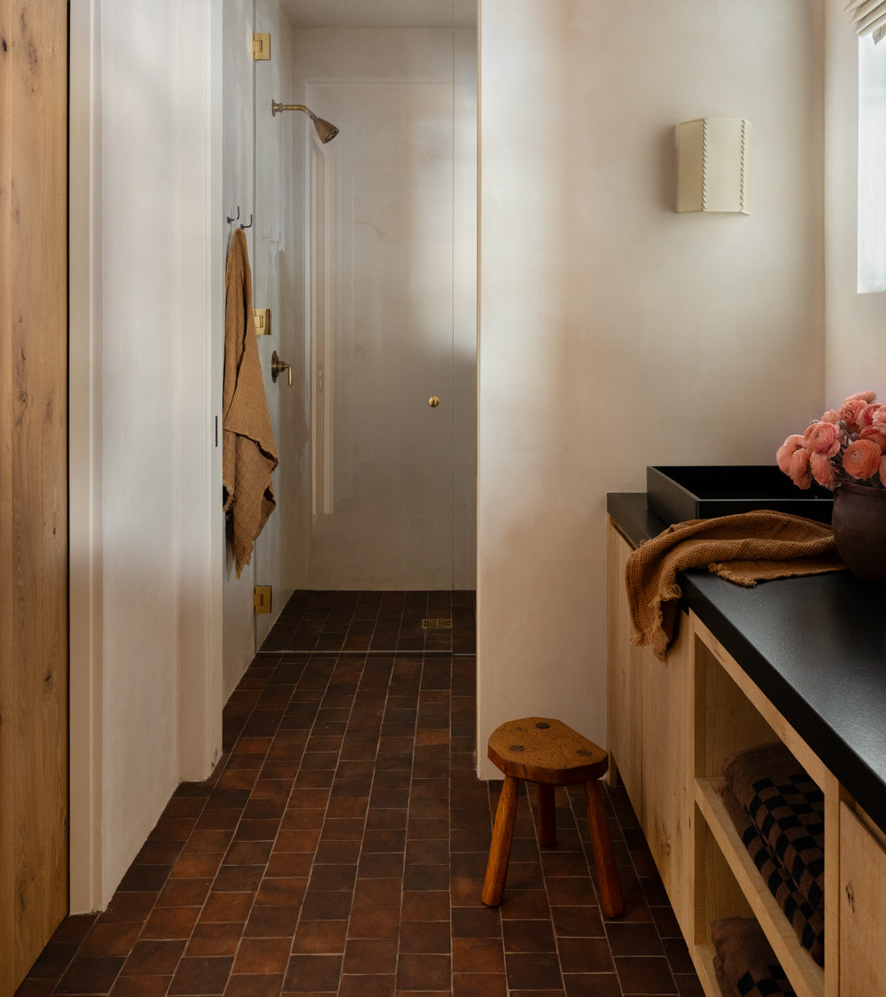 A modern bathroom featuring Zia Tile’s 4x4 Square + Madera handmade Cotto floor tiles, a walk-in shower with gold fixtures, beige walls, wooden cabinets, open shelves with towels, a black countertop sink, a stool, and pink flowers in a vase.