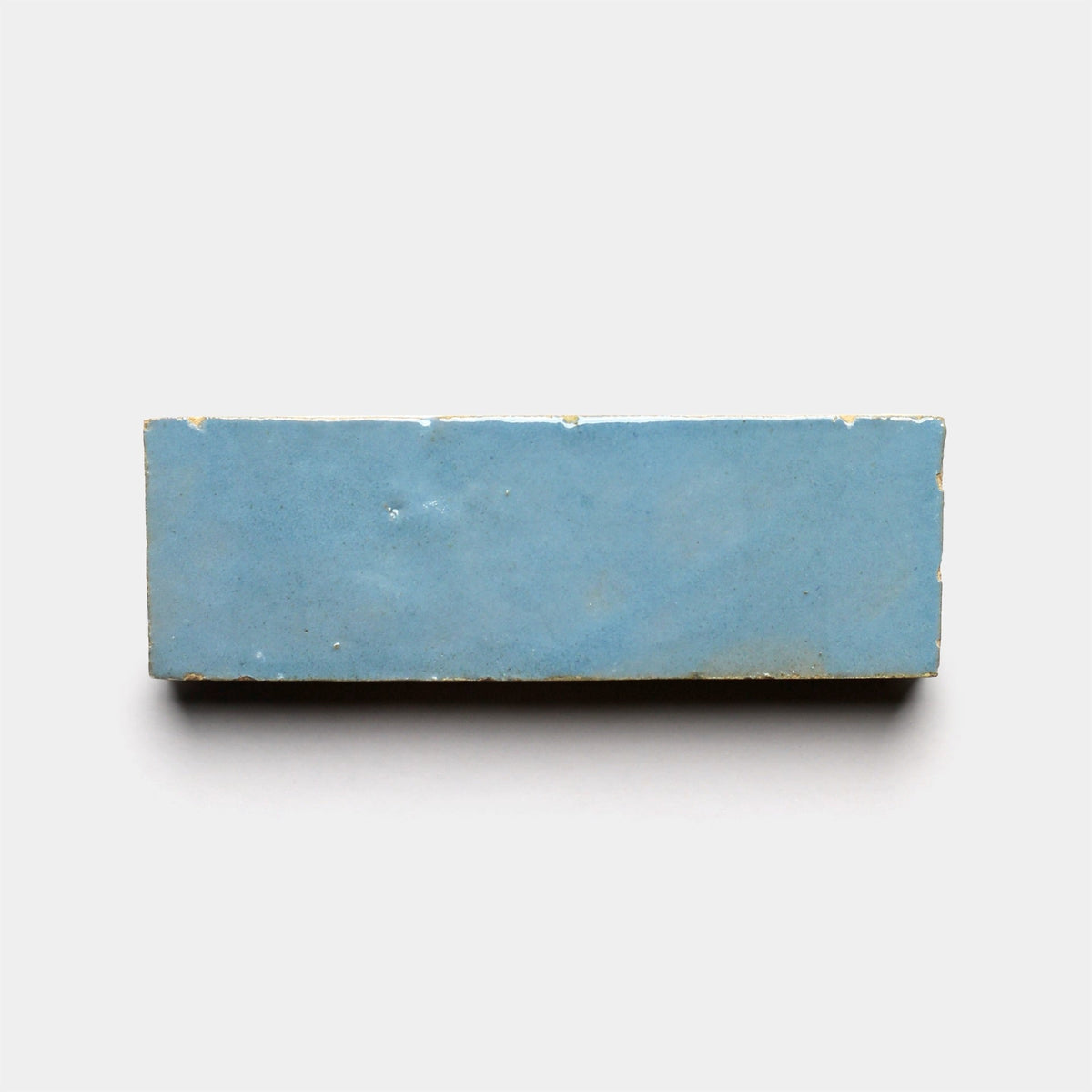 A rectangular Superior Blue 2x6 ceramic sample from Zia Tile is shown against a plain white background, featuring a smooth pale blue surface with subtle imperfections and worn edges that evoke the look of handmade tiles.