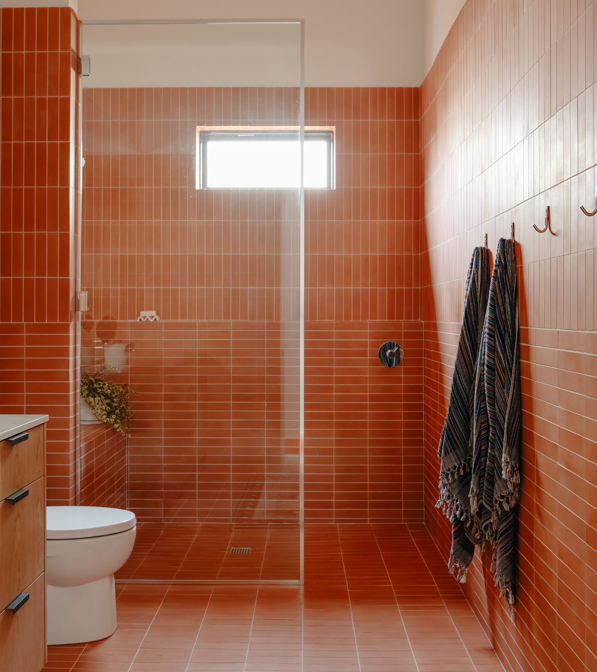Modern bathroom with Terra Cotta 2x8 tile by vendor-unknown, glass shower divider, white toilet, small window, two striped towels on wall hooks, and a plant near the shower for greenery.