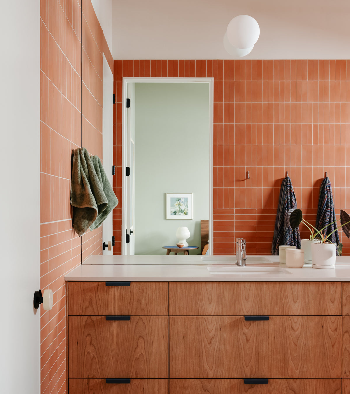 A modern bathroom features vendor-unknown Terra Cotta 2x8 subway tile, a wooden vanity with black handles, a countertop with two potted plants, hanging towels, and a mirror reflecting a green-walled room with framed art.