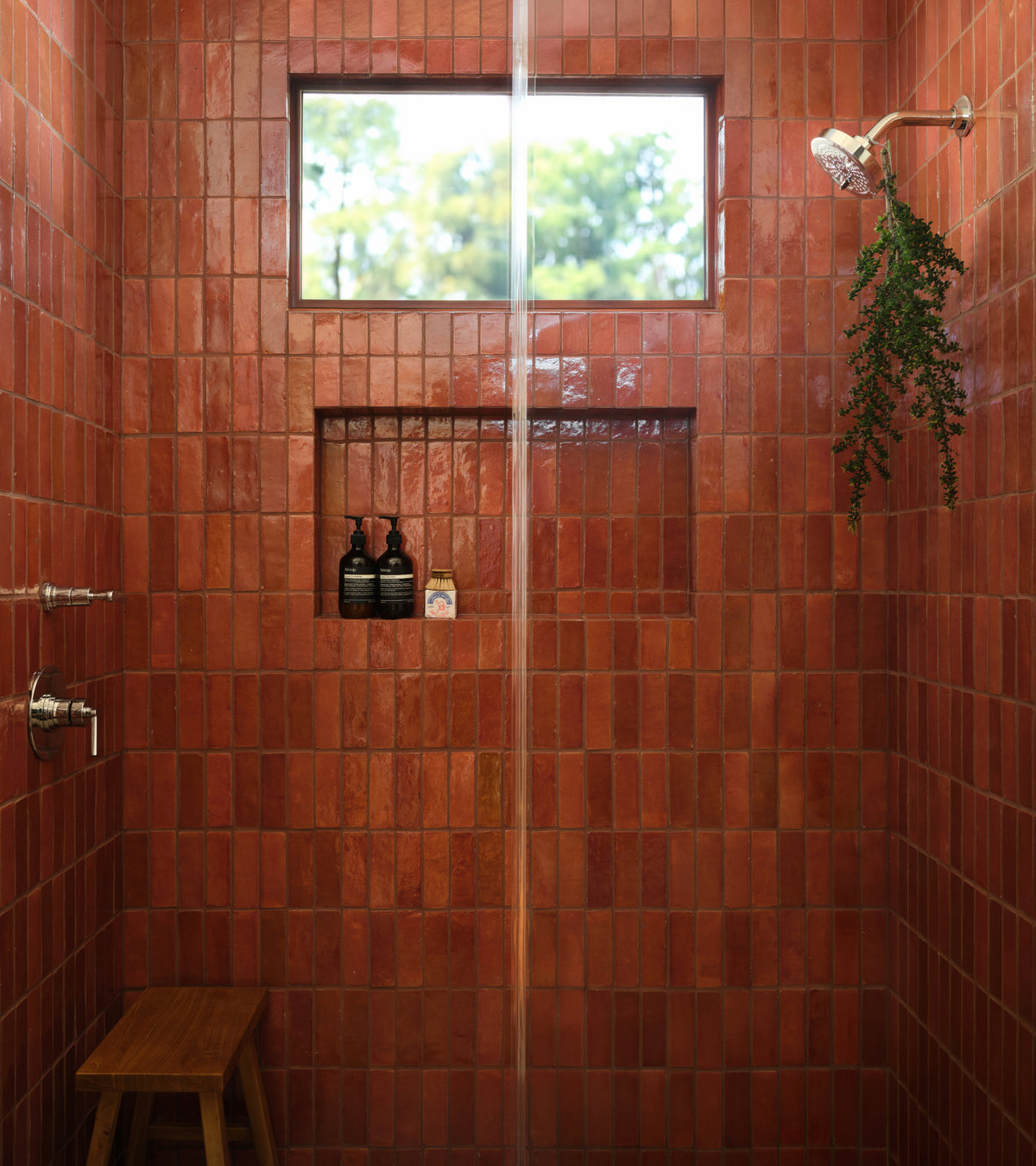 A shower stall with Terra Rosa 2x6 zellige tiles, a built-in recessed shelf holding soap bottles, a small window, and a wooden stool.