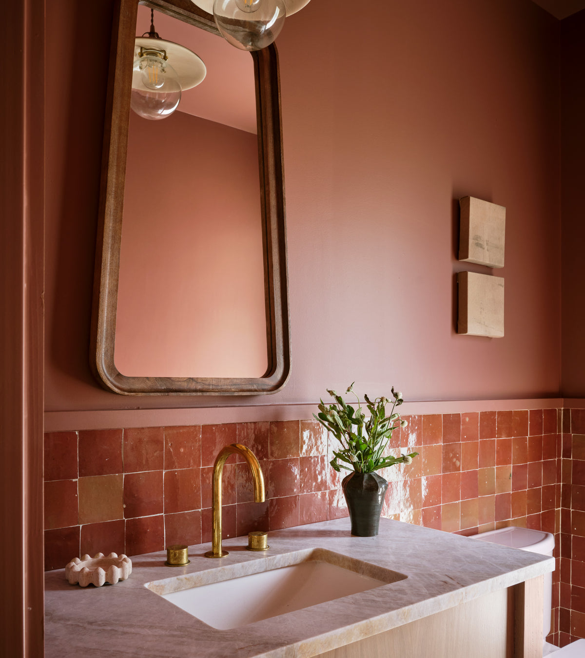 A bathroom features pinkish-brown walls, a large wood-framed mirror, gold faucet, white sink, Terra Rosa 4x4 tiles by vendor-unknown as the backsplash, two small wall art pieces, and a vase with flowers on the counter.