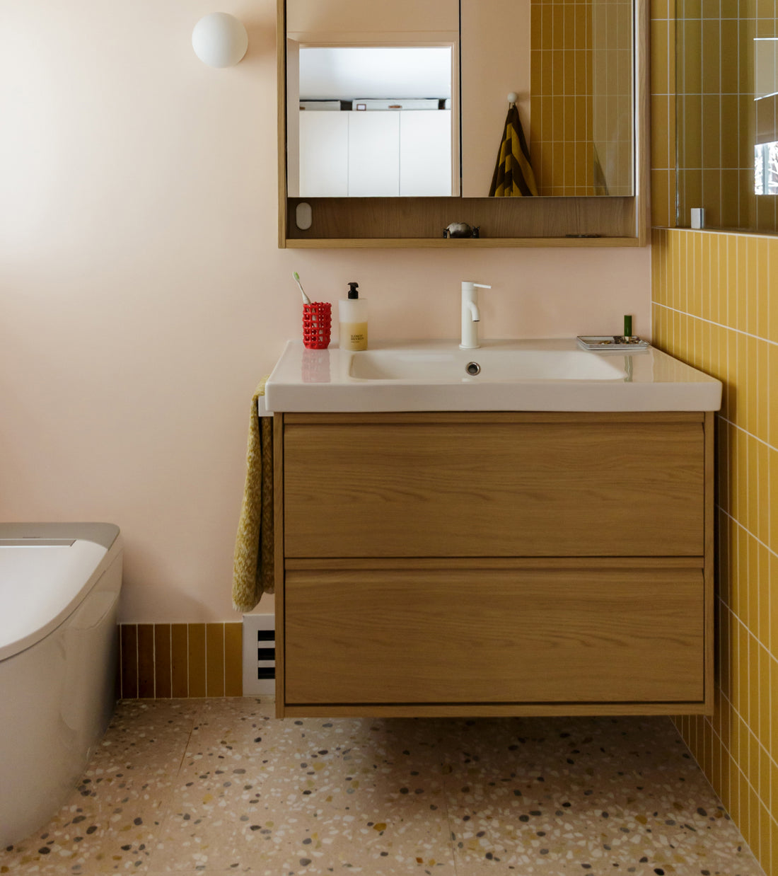 A wall-mounted wooden bathroom vanity with a white sink, set against a wall featuring yellow vertical Sunnylands 24x24 Zia Tile.