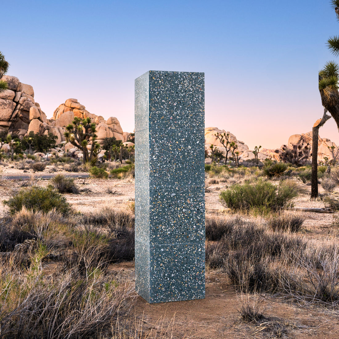 A tall, rectangular monolith stands in a desert with dry grass, rocks, bushes, and Joshua trees at sunset—its surface gleams like Thunderbird 24x24 handmade tile from Zia Tile.