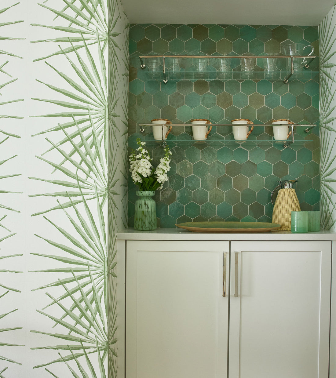 White cabinet with two doors, Zia Tile’s Tidepool Hex green Moroccan tile backsplash, glass shelf with glassware, three white mugs hanging, green vase with white flowers, yellow textured container, and leafy side-wall wallpaper.