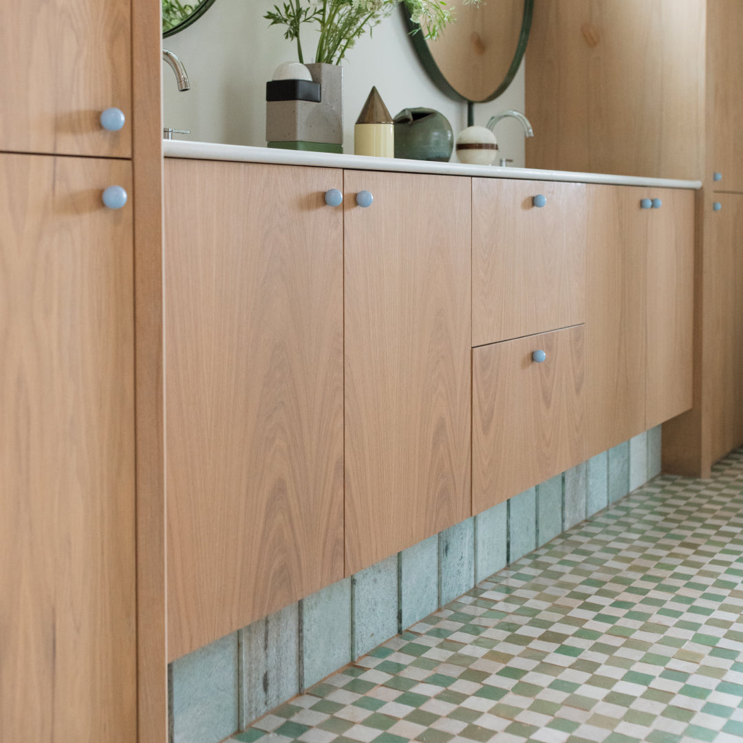 Light wood bathroom vanity with blue knobs featuring Tidepool + Casablanca Checkerboard handmade Moroccan tiles on the floor and sage green tiled base.