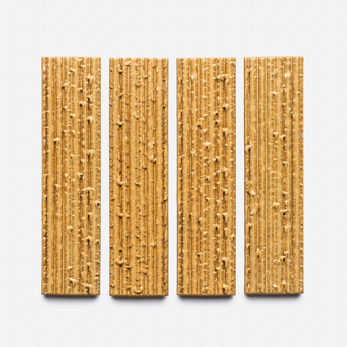 Four Toast + Furrowed, Sample crackers by Zia Tile, rectangular with vertical ridges and a golden-brown texture, are evenly spaced in a row on a plain white background.
