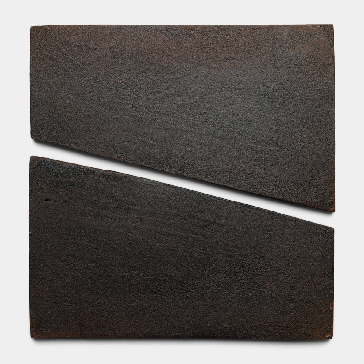 Two rectangular, dark, textured Toltec + Oscura tiles by Zia Tile are placed side by side, with one slightly angled so their inner edges misalign, creating a diagonal gap between them.