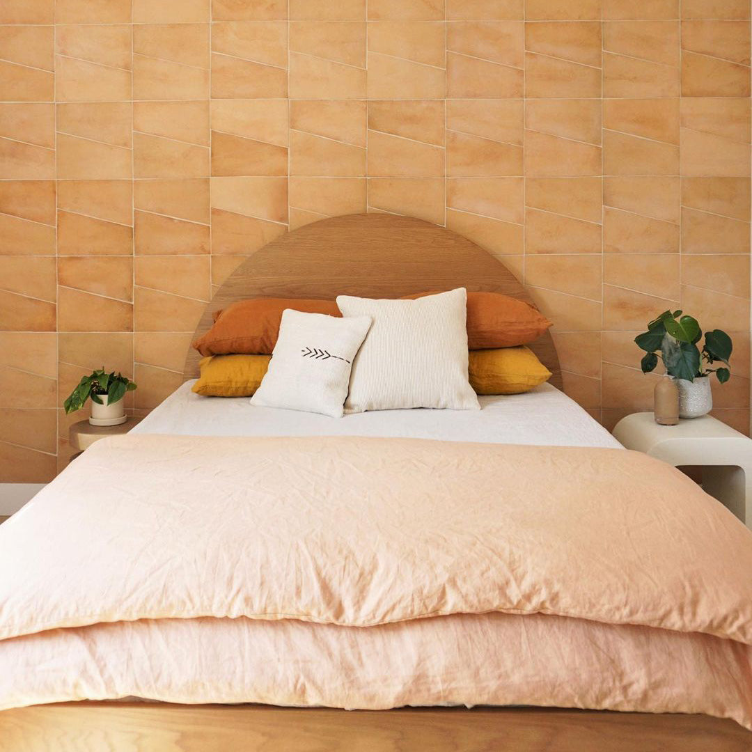 A neatly made bed with peach and white bedding, two orange and two white pillows, against a tiled peach-colored accent wall. Small plants sit on bedside tables on each side of the bed.