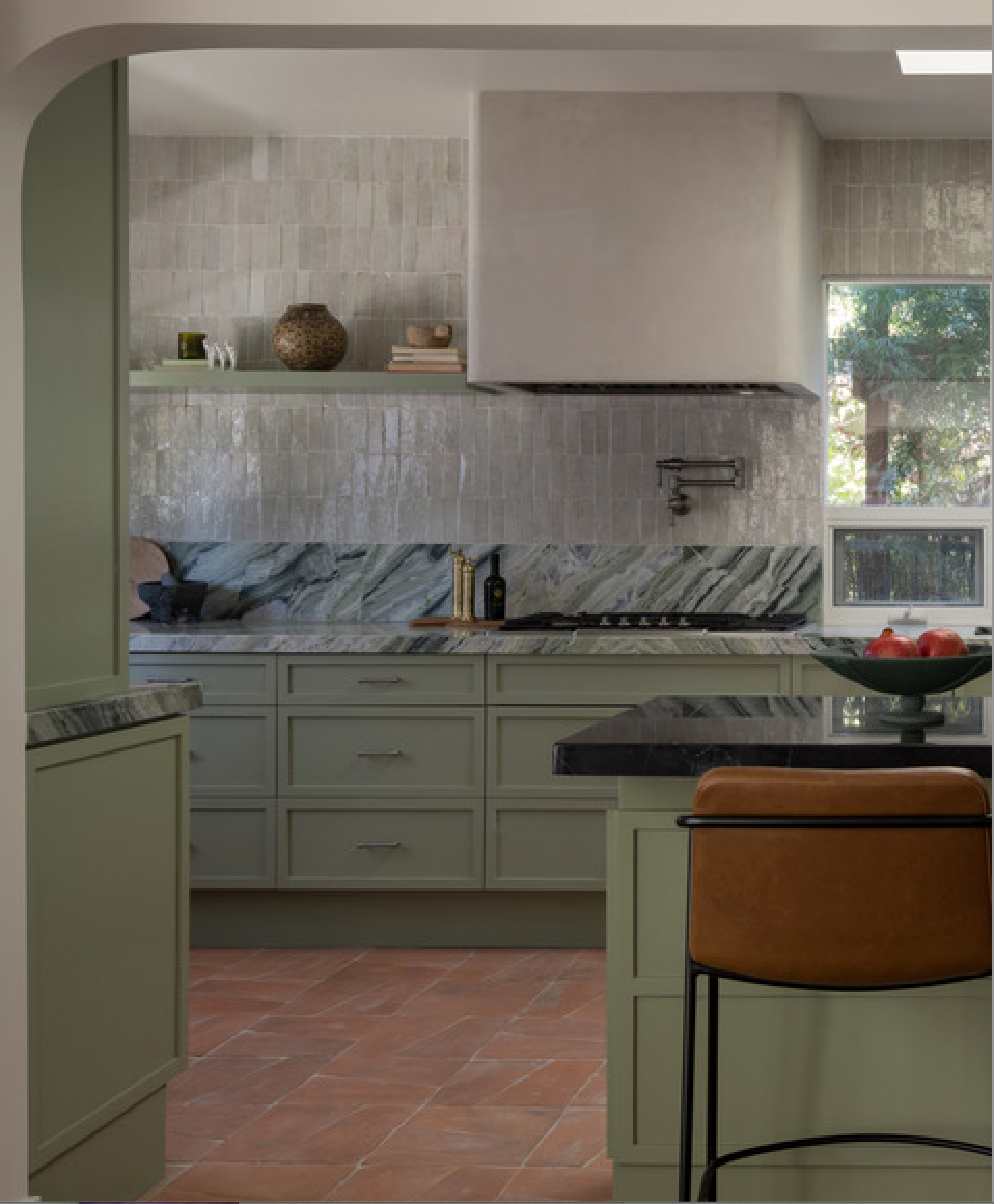 A modern kitchen with green cabinets, marble backsplash, and terracotta floor tiles. There is a black countertop with a bowl of red apples, a brown leather barstool, and textured neutral tiles on the walls.