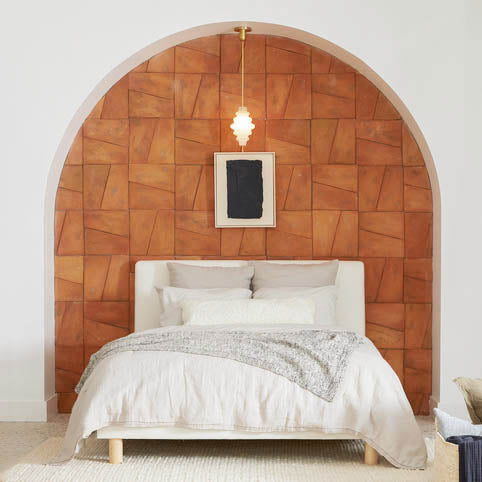 A cozy bedroom with a white bed and neutral bedding is set against a terracotta geometric accent wall within an arched alcove. A small pendant light and minimalist framed artwork hang above the bed.
