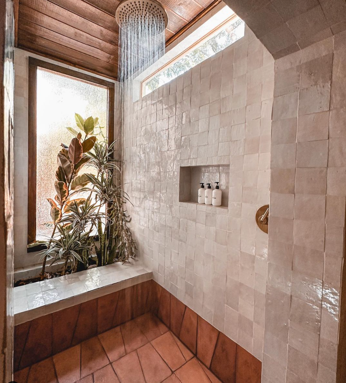 A bright, modern shower with white tile walls, a built-in bench, large window, indoor plants, and rainfall showerhead. Natural light streams in, highlighting shampoo bottles in a recessed shelf.