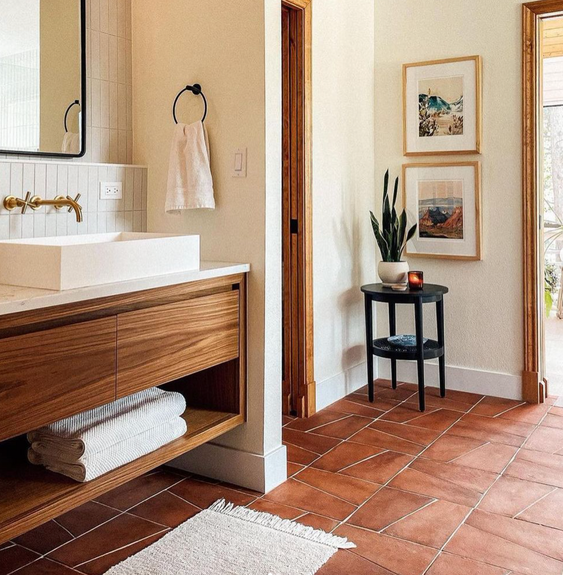 Bathroom with terracotta tile floor, wooden vanity with white sink, towel ring, white towel, wall mirror, black side table with plant, candle, two framed prints, and natural light coming from a doorway.