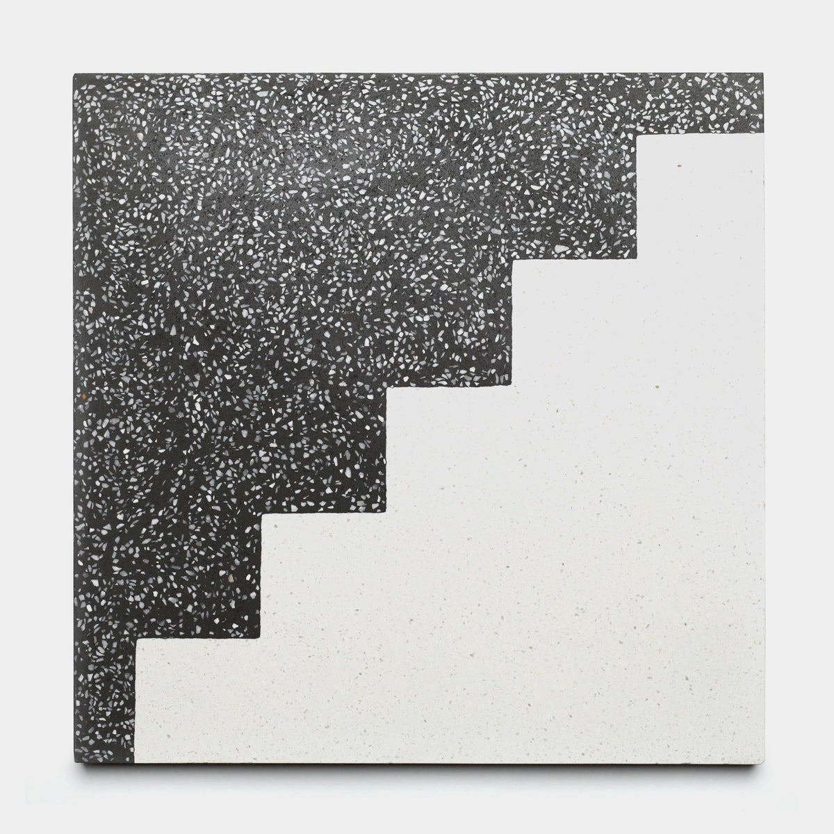 The Zia Tile Trellick Ash 12x12 sample features a black and white speckled terrazzo design, divided diagonally with a stepped zigzag line—black on the upper left, white on the lower right.