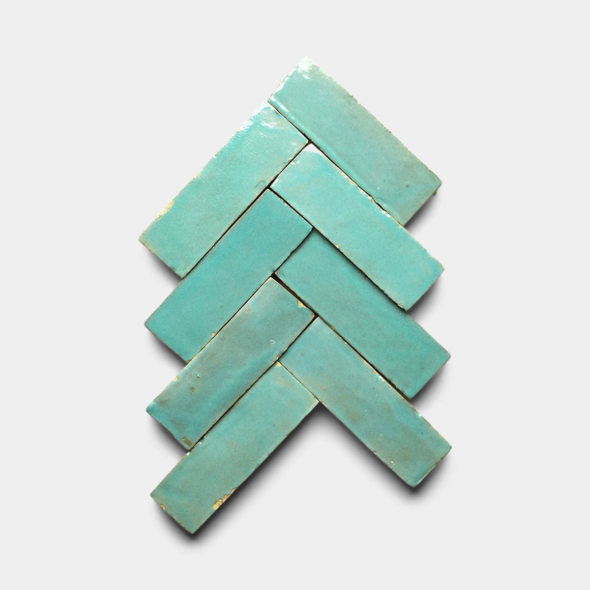 Seven Tulum 2x2 turquoise cement tiles by Zia Tile are arranged in a herringbone pattern on a light gray background, showcasing their handmade, rustic texture and subtle color variations.