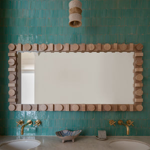 A rectangular mirror with a chunky wooden frame is mounted on a teal Zia Tile Tulum Trapezoid wall above a stone countertop with dual sinks, brass faucets, and a shell dish. A textured pendant light hangs overhead.