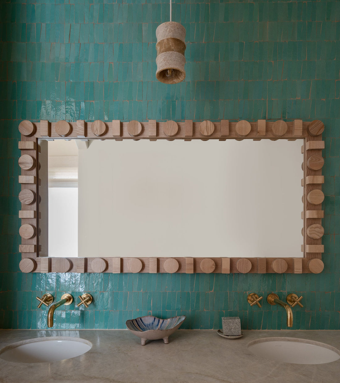 A rectangular mirror with a chunky wooden frame is mounted on a teal Zia Tile Tulum Trapezoid wall above a stone countertop with dual sinks, brass faucets, and a shell dish. A textured pendant light hangs overhead.