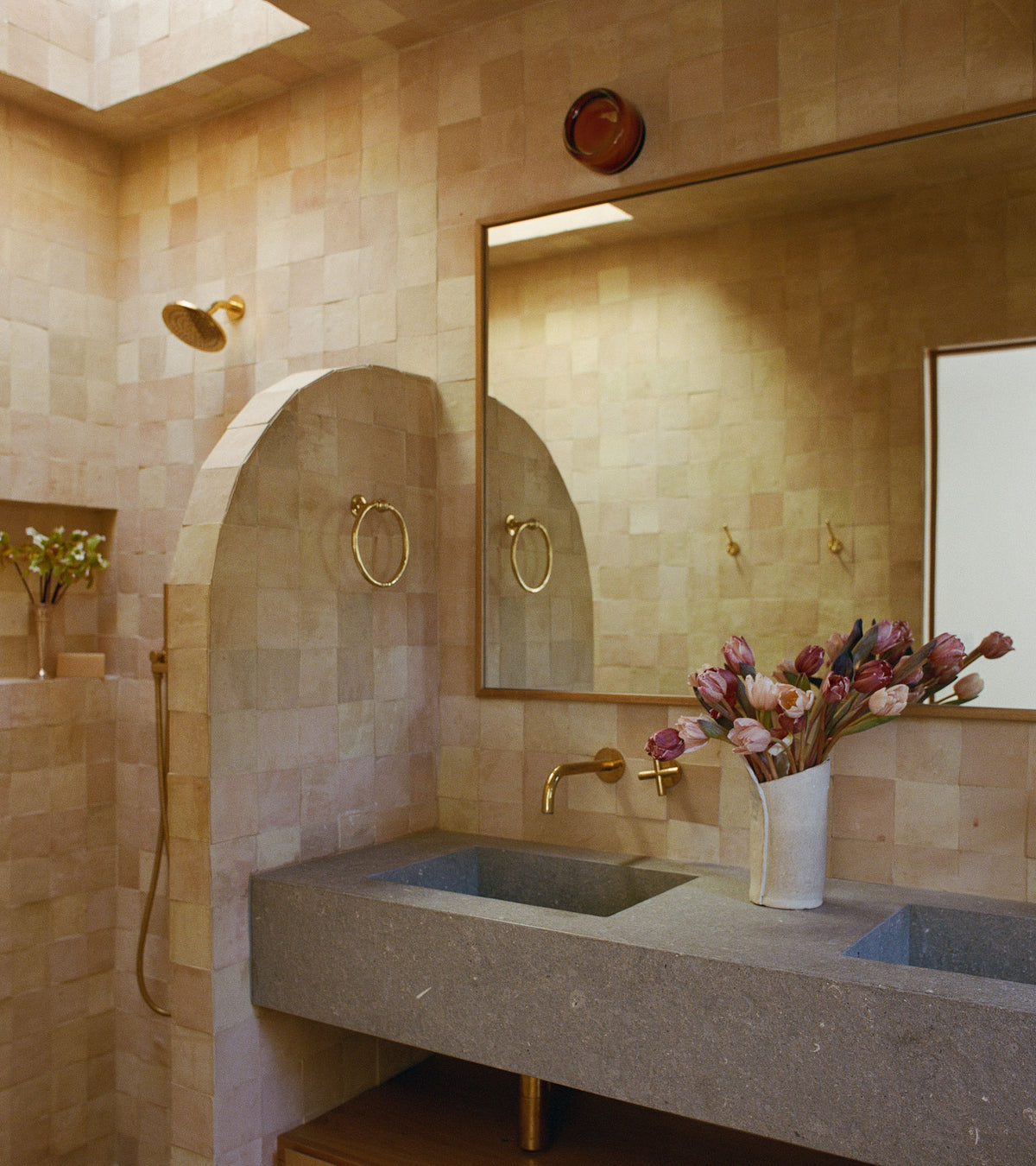 A modern bathroom with Unglazed Natural 4x4 tiles by Zia Tile on the walls, an arched shower, a large mirror, dual stone sinks, brass fixtures, pink flowers in a vase, and skylight streaming in natural light.