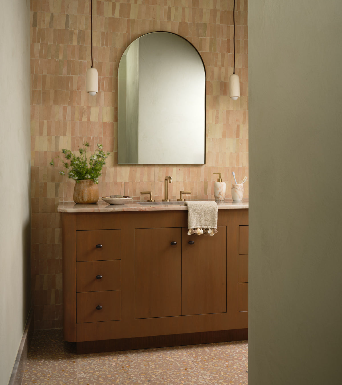 A modern bathroom features a wooden vanity, gold fixtures, a large arched mirror, and walls clad in Unglazed Natural Trapezoid tile from Zia Tile. Pendant lights frame the mirror, while a small plant and towel rest on the countertop.