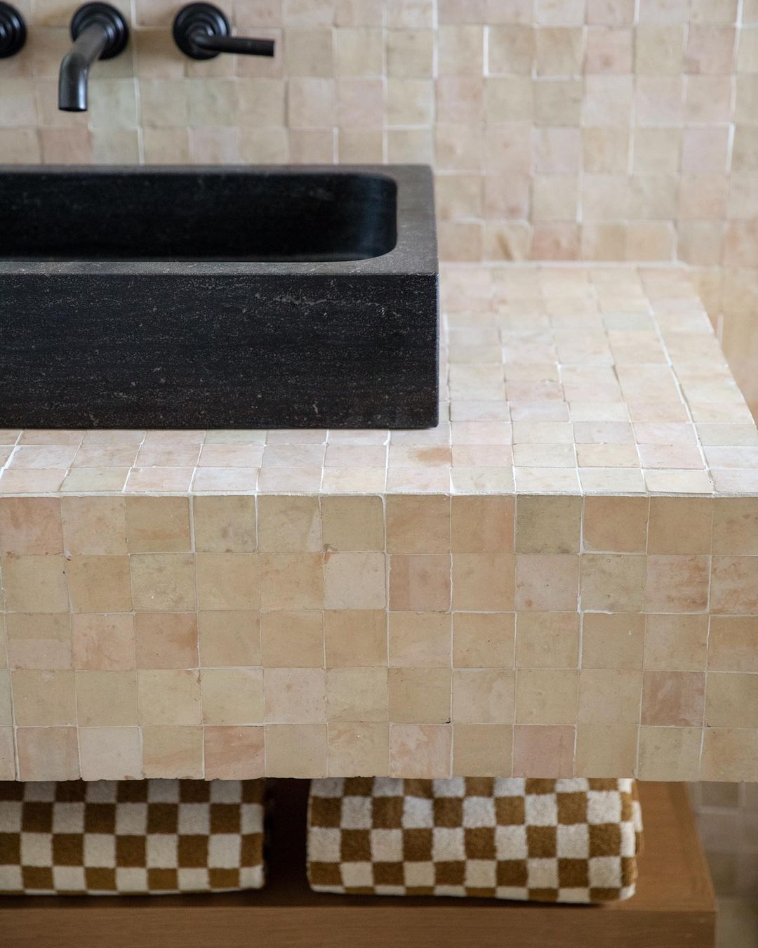 A black rectangular sink with a matte black faucet sits on a countertop covered in Zia Tile’s Unglazed Natural 2x2 tiles; two brown and white checkered baskets are visible on the shelf below.