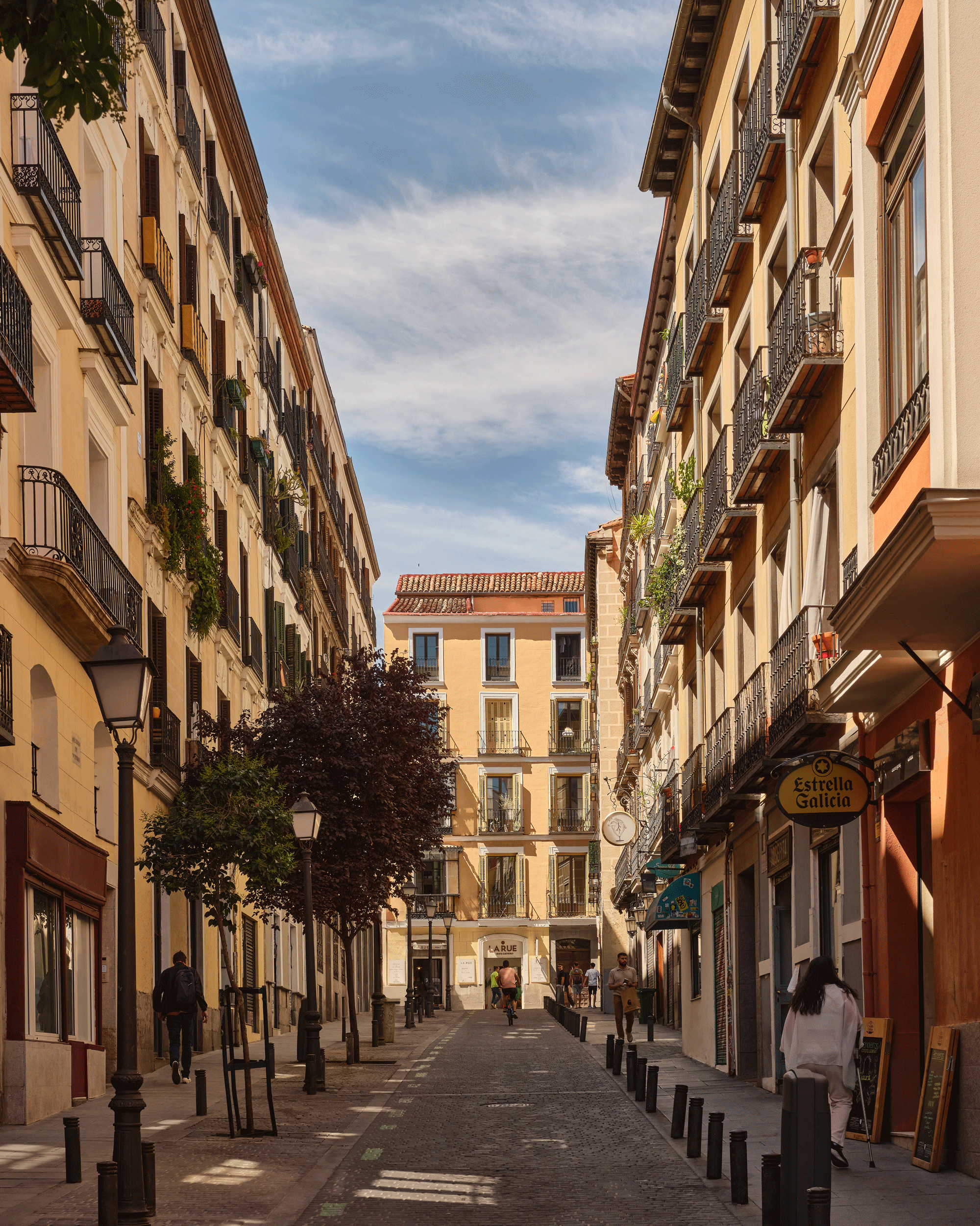 A narrow European street lined with tall, colorful buildings featuring balconies and plants. People walk along the cobblestone road beneath a partly cloudy sky. Shops and streetlights are visible on both sides.