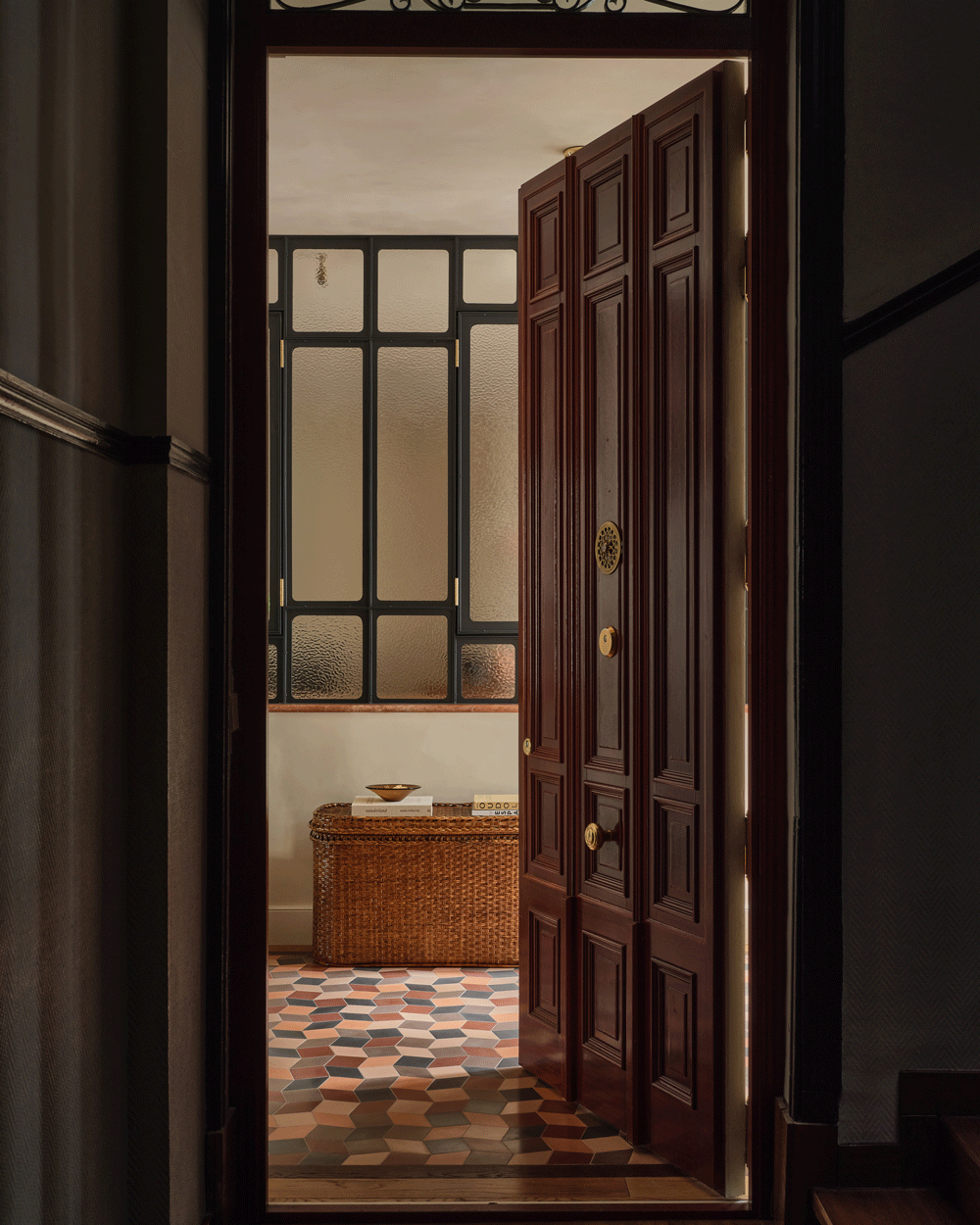 A dark wooden door stands ajar, revealing a hallway with patterned tile flooring, frosted glass windows with black frames, and a woven basket bench against the wall. Warm, soft lighting fills the space.