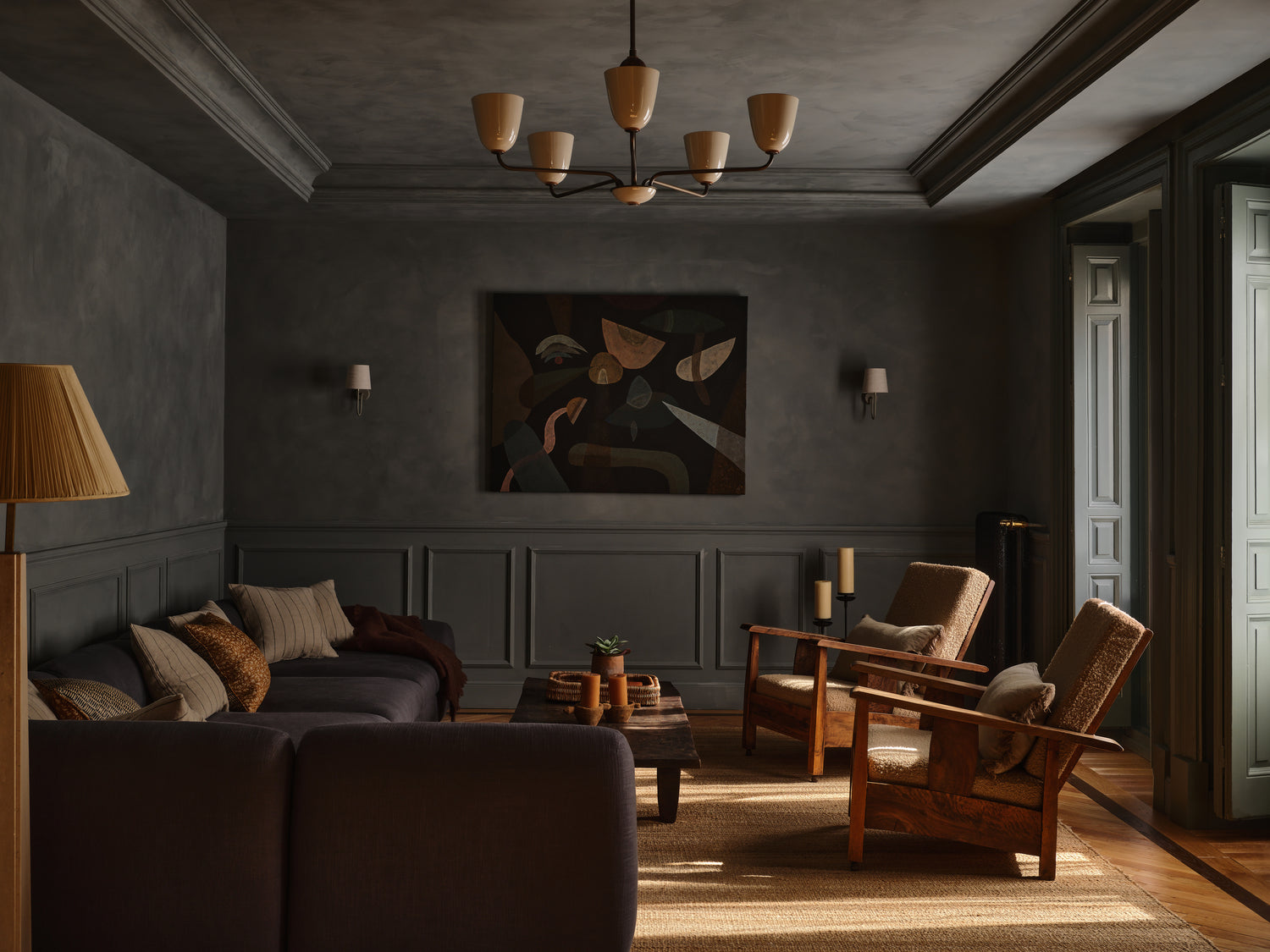 A cozy, dimly lit living room with dark gray walls, wooden furniture, a sectional sofa with cushions, two armchairs, a wooden coffee table, abstract wall art, and large windows letting in natural light.