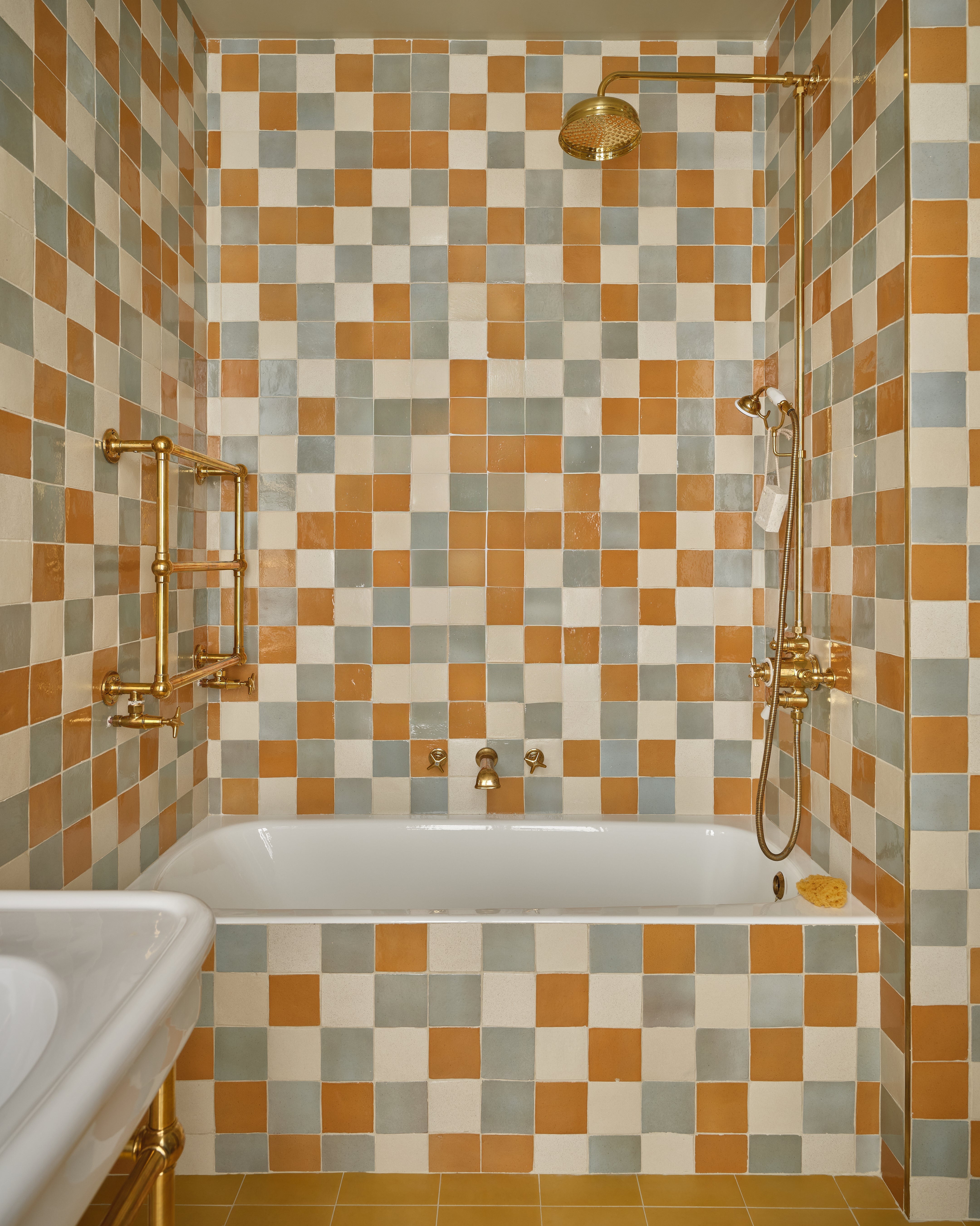 A bathroom with a bathtub and a shower, surrounded by square tiles in orange, gray, and cream colors. The fixtures, including the showerhead, faucet, and towel rack, are gold-toned.