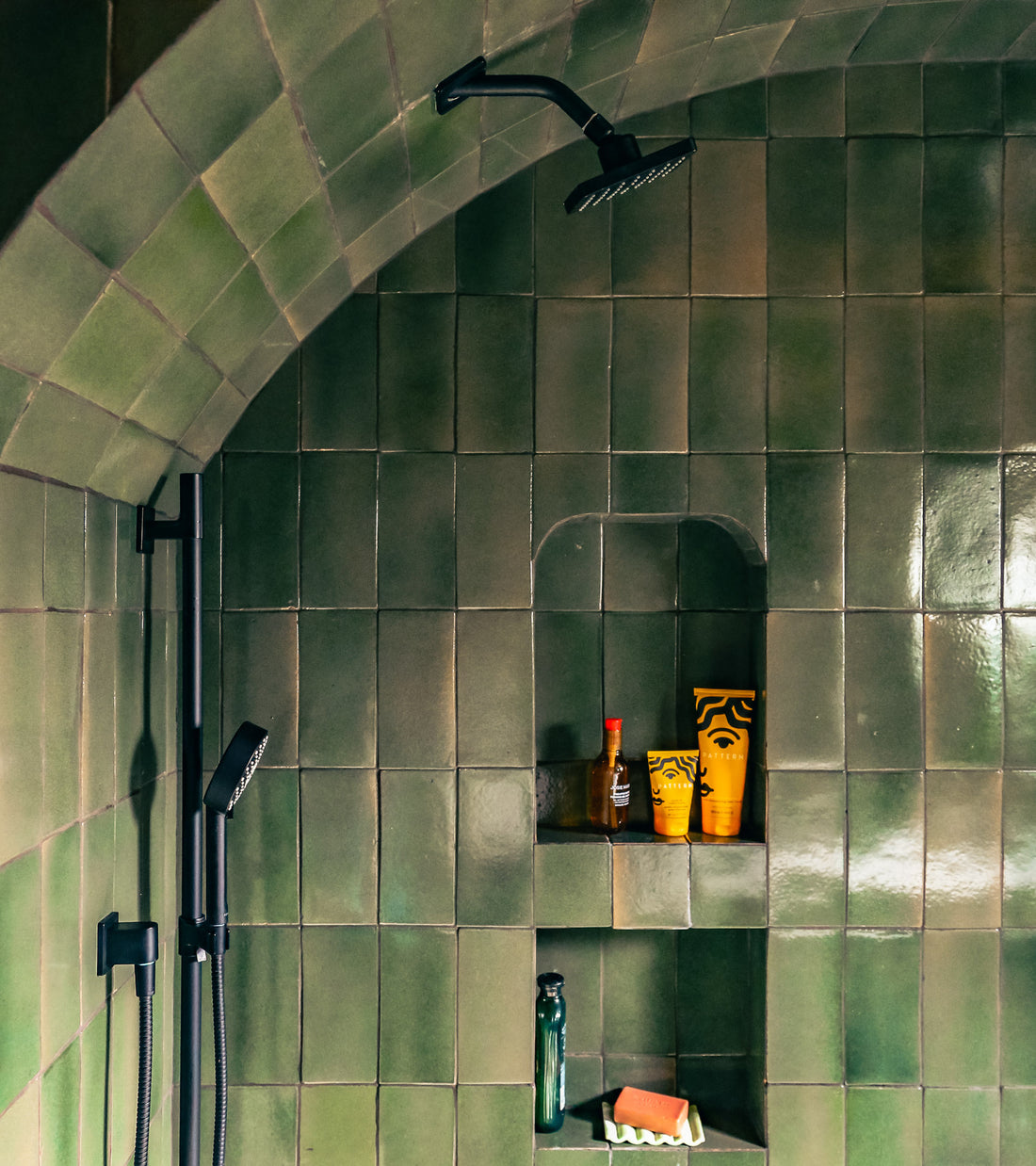 A shower area with Zia Tile Valverde 4x8 Rectangle green tiles, a black overhead and hand shower, plus built-in shelves holding toiletries: a Tabasco bottle, two yellow tubes, soap bar, and a dark green bottle.