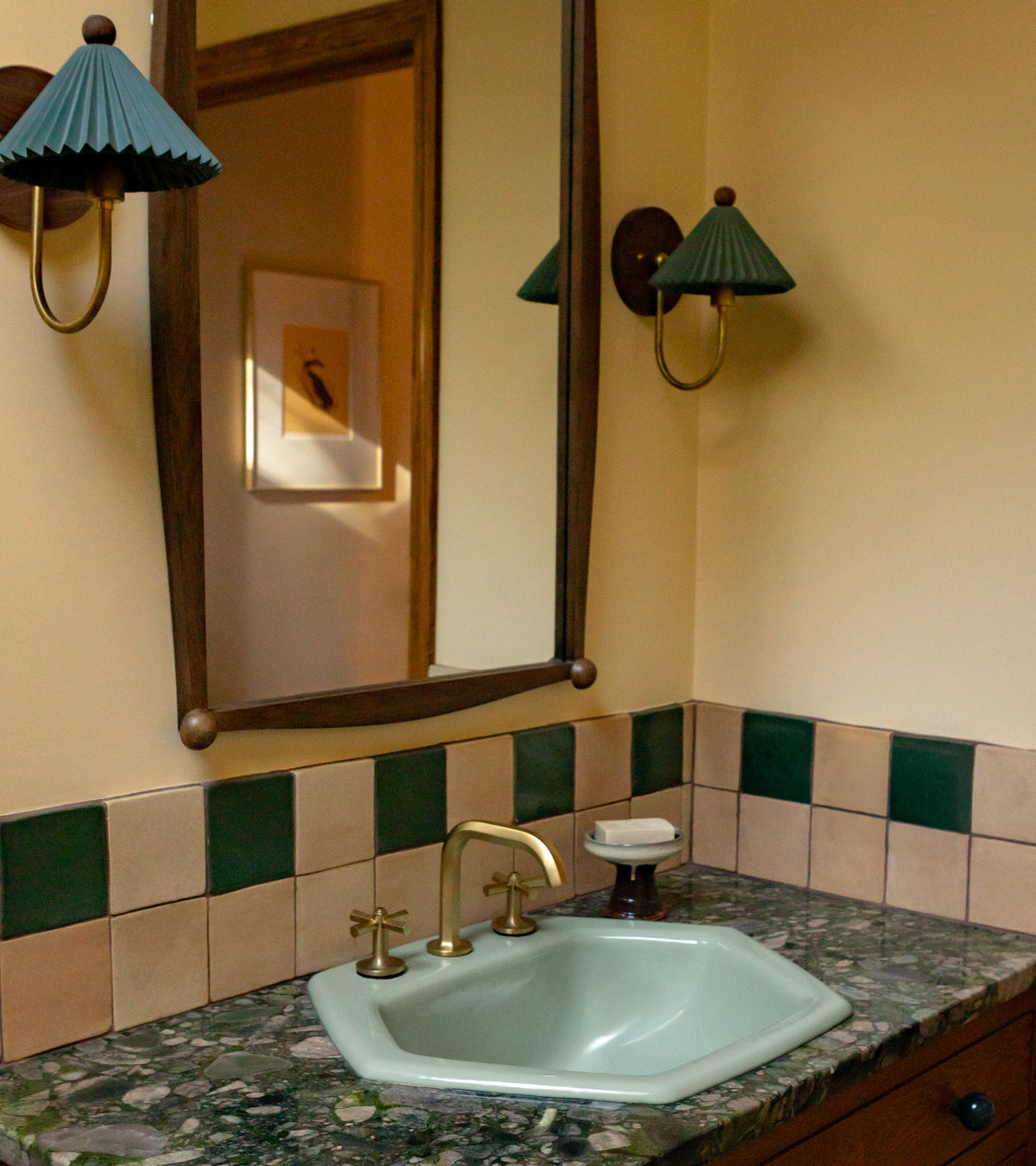 A bathroom vanity features a green marble countertop, pale green sink, brass faucet, and a handmade Arroyo 4x4 Square backsplash by Zia Tile. Above the sink are a large mirror and two wall sconces with green shades.