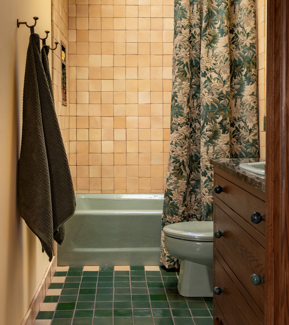 A small bathroom with Zia Tile Valverde 4x4 Square beige tiles on the walls, handmade dark green and cream checkered Cotto floor tiles, a light green bathtub, floral shower curtain, toilet, dark towels on hooks, and a wooden vanity.