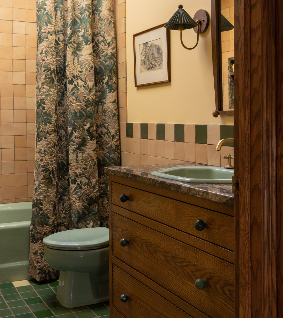 A vintage-style bathroom with green fixtures, a wood vanity, marble countertop, Zia Tile’s Valverde 4x4 Square tiles in green and tan, a leafy shower curtain, wall-mounted light, and framed art above the sink.