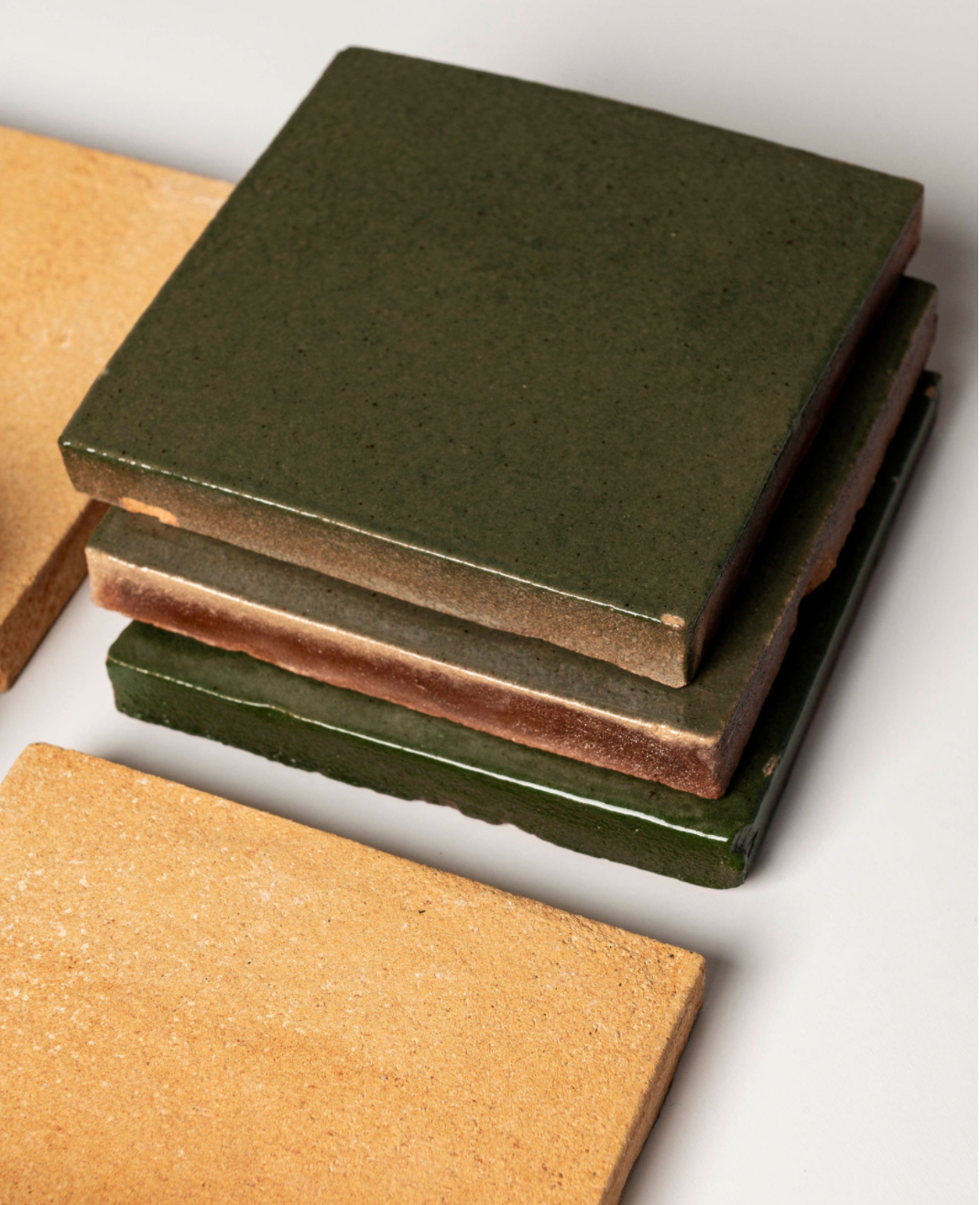 A stack of Zia Tile Valverde 4x4 Square handmade cotto tiles in earthy dark green, brown, and ochre tones sits on a light background with two ochre tiles lying flat beside the stack.