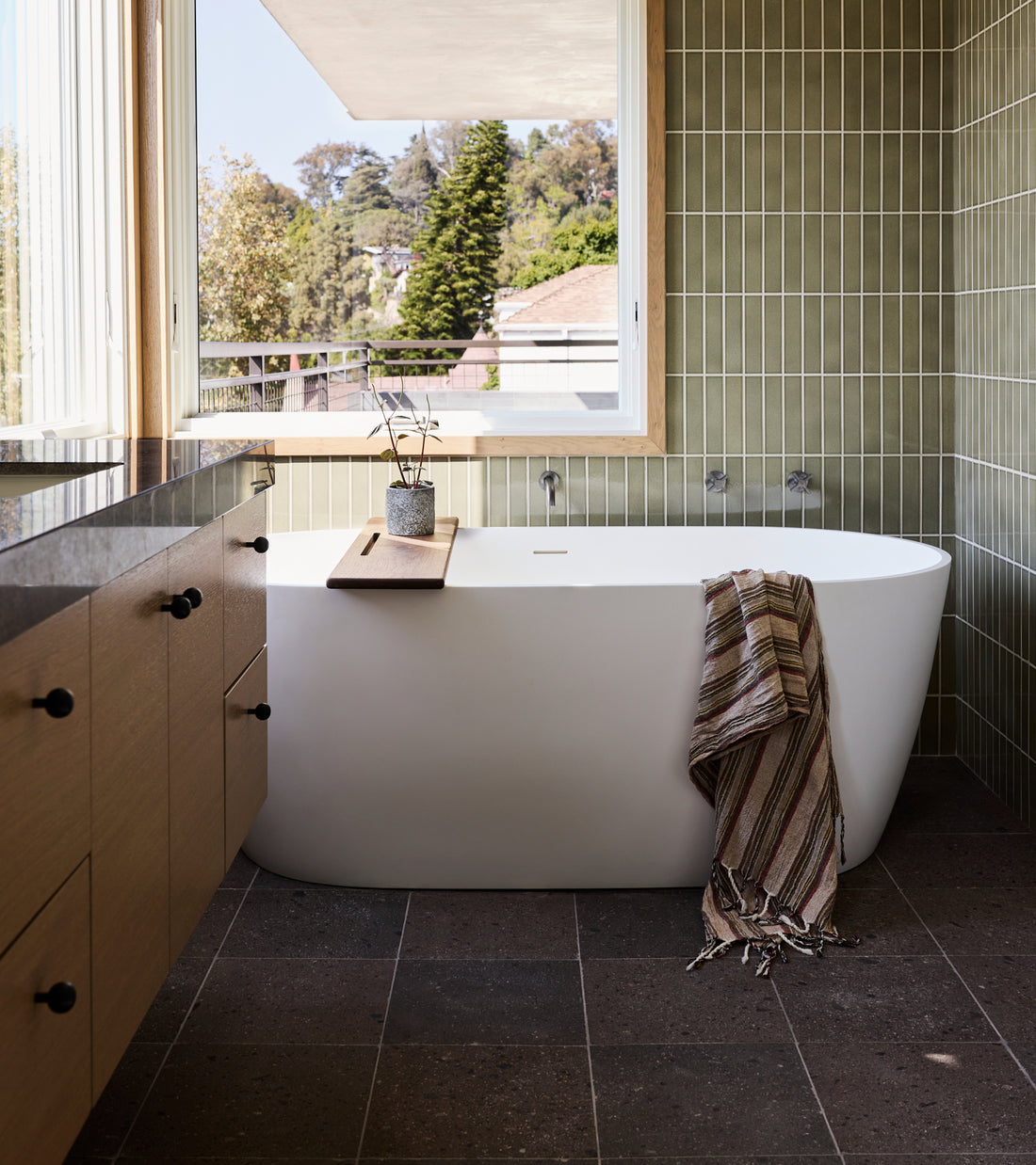 A modern bathroom with a freestanding white tub, a wooden tray holding a small plant, and green vertical Volcan 12x12 Mexican Cantera tile walls by Zia Tile.