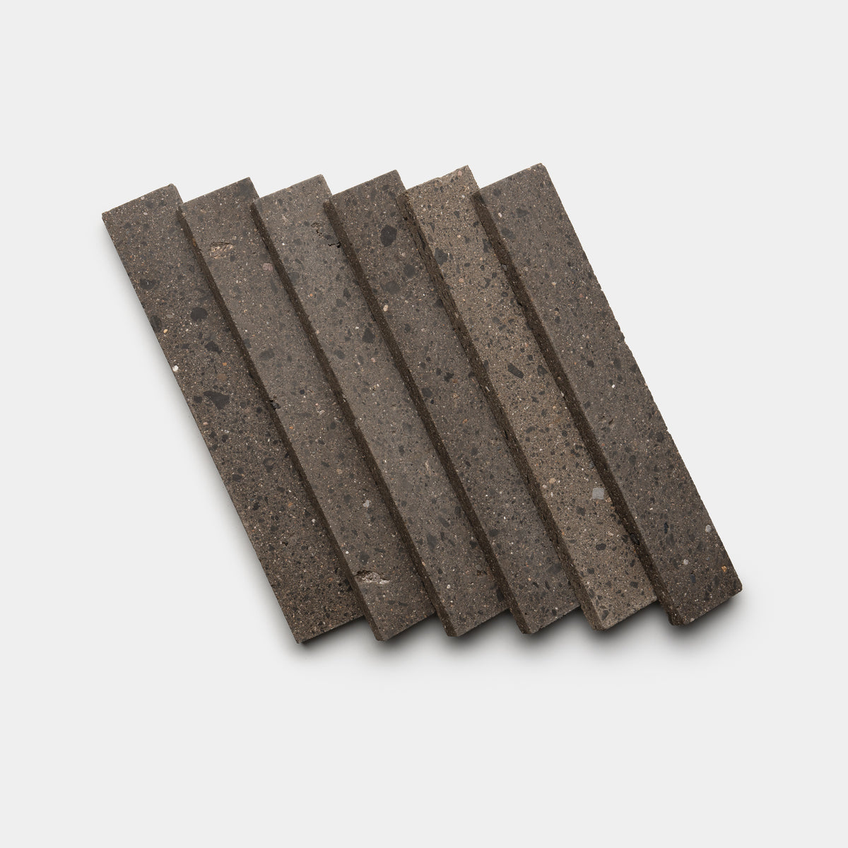 Six Volcan 2x12 tiles by Zia Tile, in dark gray stone, are arranged diagonally on a white background with slight overlap in a neat, evenly spaced formation.
