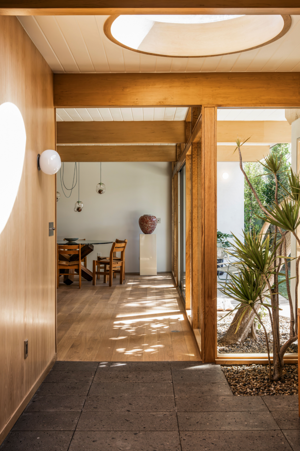 A modern interior with wood beams, large windows, and a sunlit dining area. Indoor plants grow in a small rock garden, and natural light highlights the open, minimalist space.