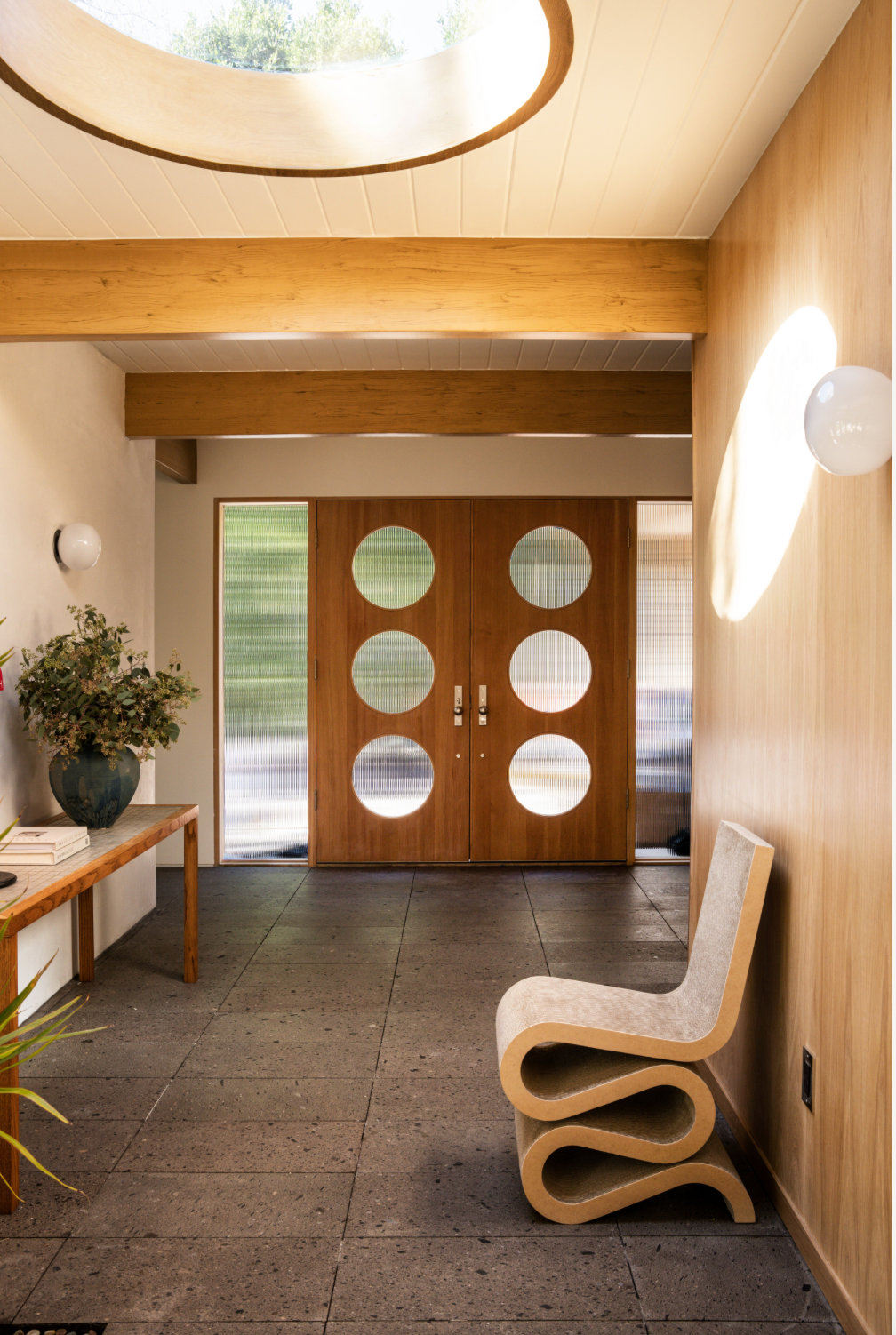 A modern entryway with wooden double doors featuring six large circular windows, a sculptural beige chair, a console table with a vase of greenery, and natural light streaming through a round ceiling opening.