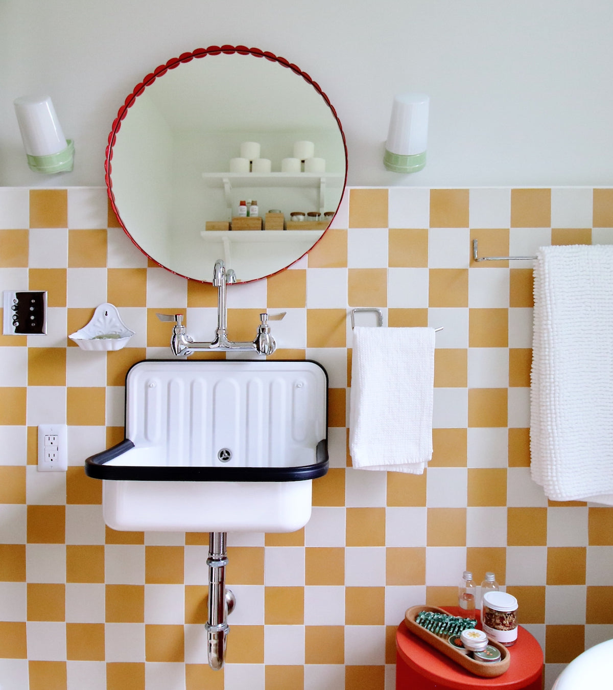 A bathroom featuring White 4x4 tiles by vendor-unknown in a yellow and white checkered pattern, a wall-mounted sink with chrome faucet, round red-framed mirror, white towels, shelves for toiletries, and a small red side table.