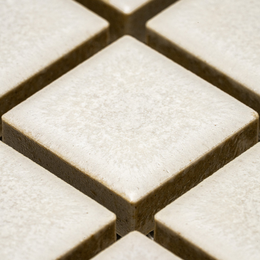 Close-up view of Zia Tile's Wool 1x1 beige ceramic tiles, arranged diagonally in a grid pattern with visible grout lines. The tiles feature a slightly textured surface.