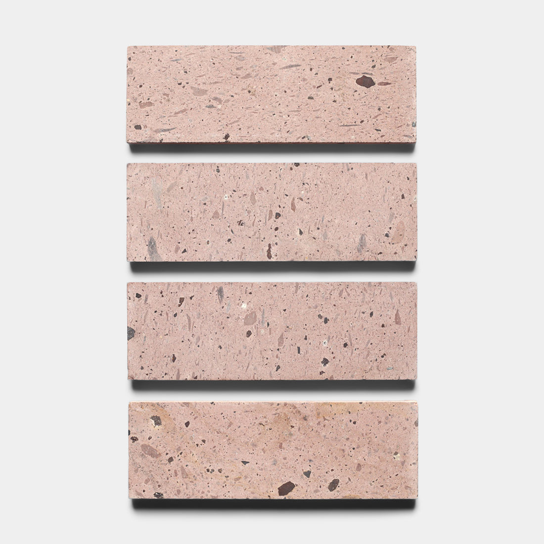 Four Yuma 4x12 tiles by Zia Tile, featuring a light pink terrazzo base with dark and light specks, are arranged vertically against a white background, reminiscent of classic Cantera stone.