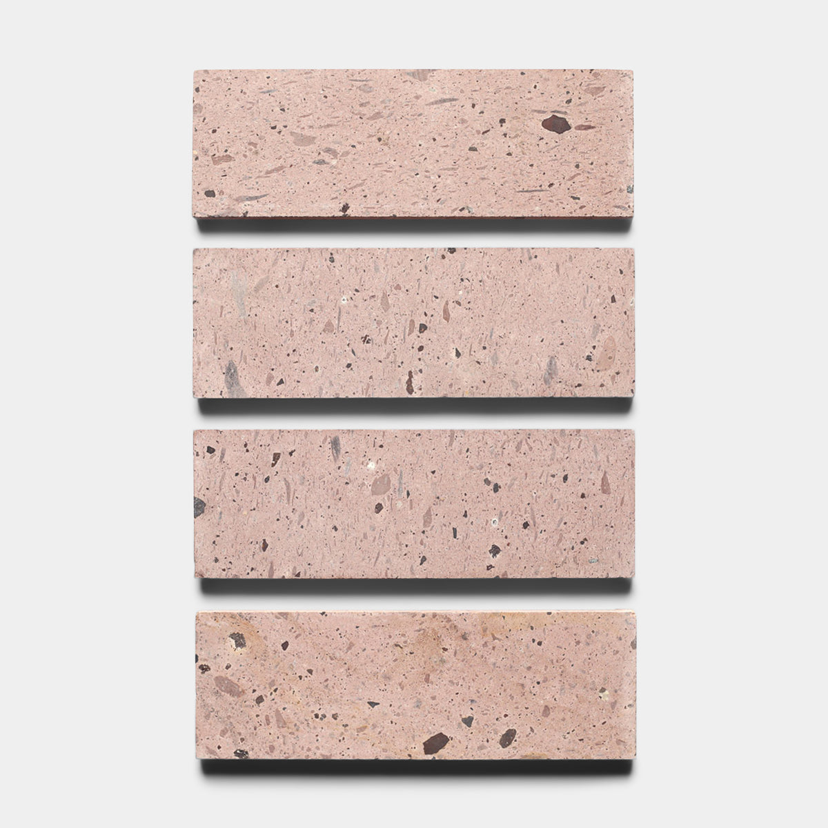 Four Yuma 4x12 tiles by Zia Tile, featuring a light pink terrazzo base with dark and light specks, are arranged vertically against a white background, reminiscent of classic Cantera stone.