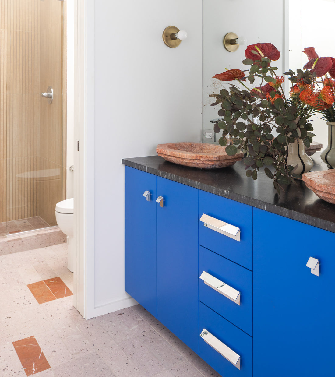 A modern bathroom features bright blue cabinets, a black countertop, large mirror, leafy vases, and Zia Tile’s Yuma 4x12 volcanic stone tile flooring near the shower and toilet in the background.
