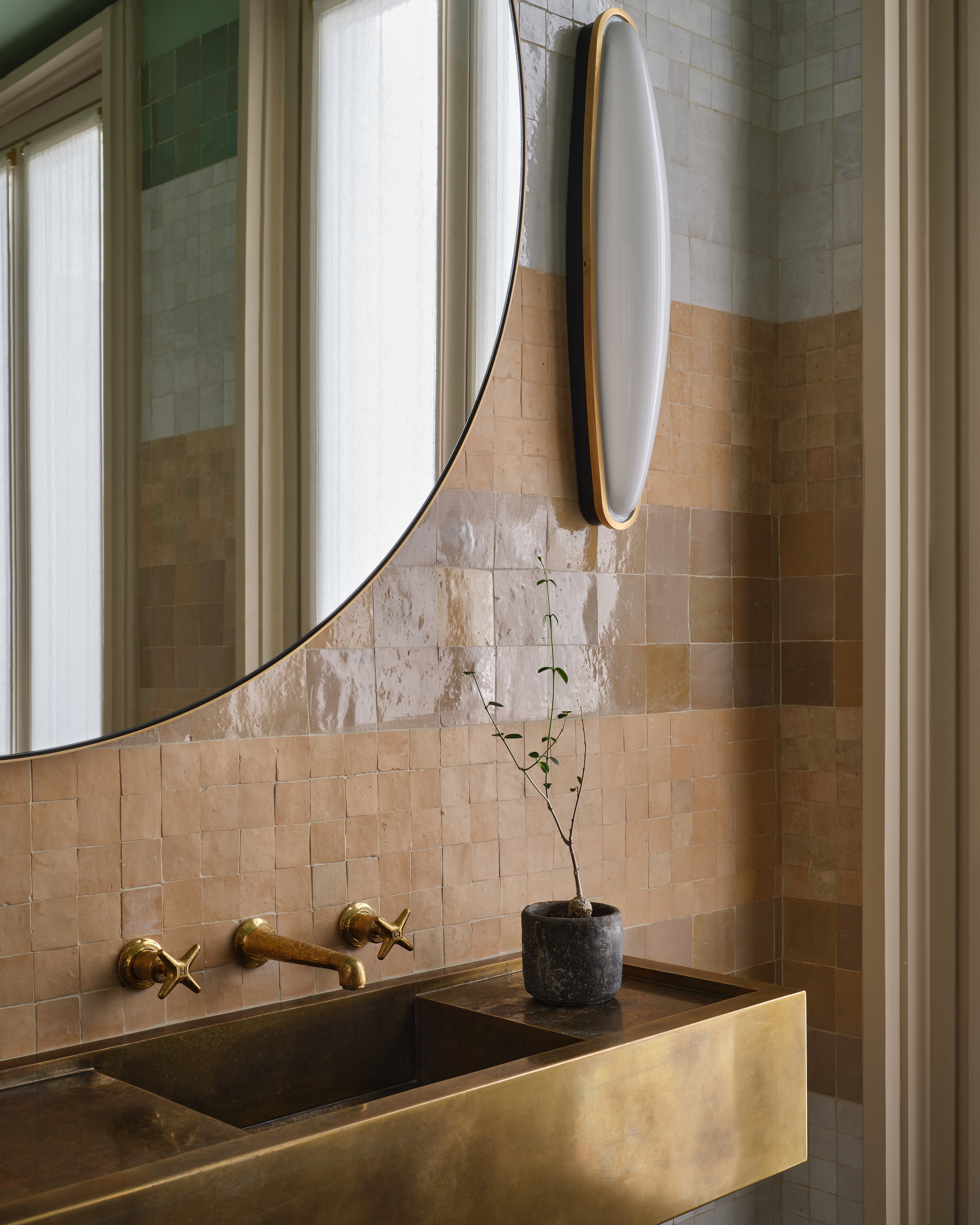 A modern bathroom features a brass sink and faucet, a small potted plant, and square beige tiles on the wall. A large round mirror and an oval wall light hang above the sink, with natural light coming through sheer curtains.