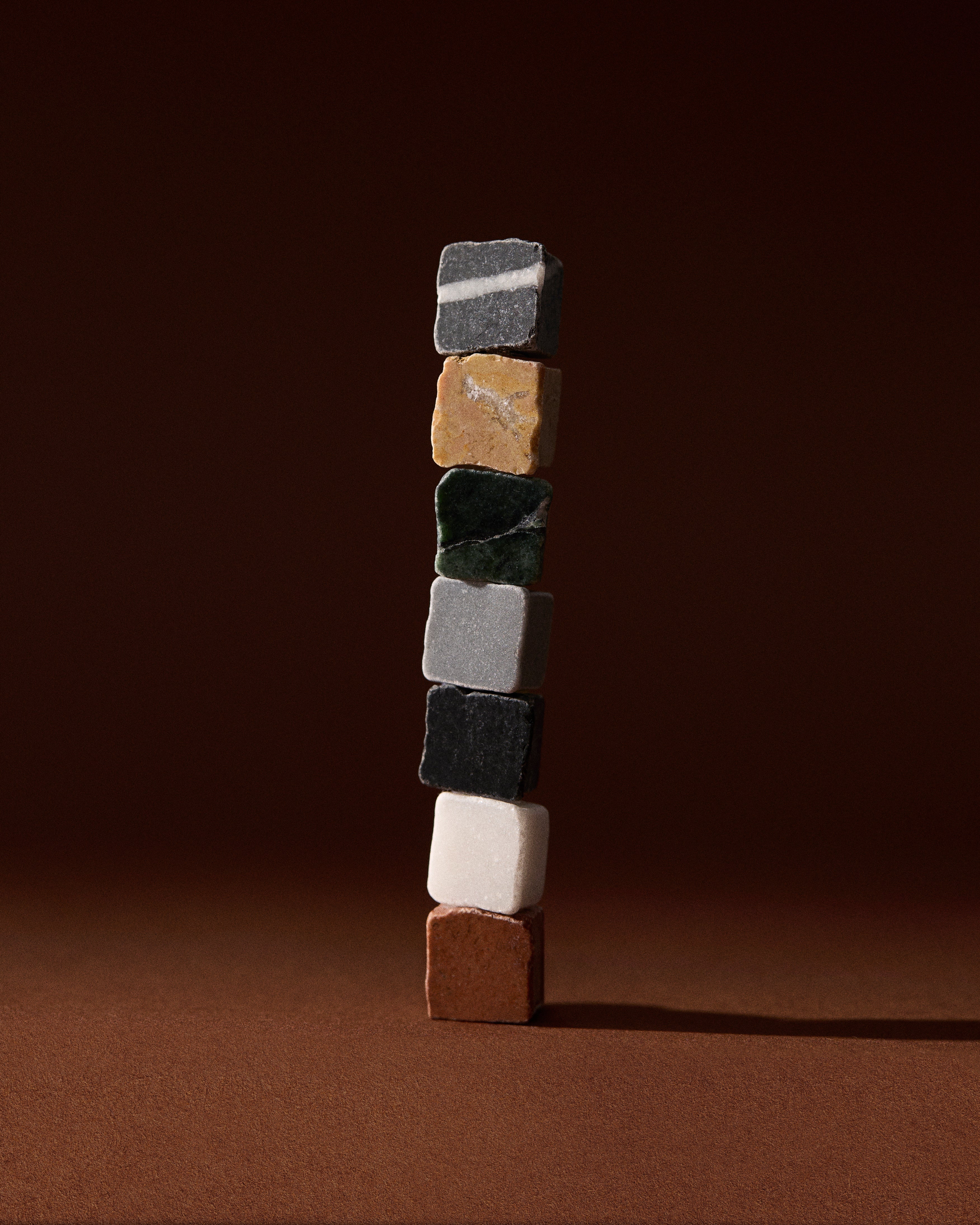 A vertical stack of seven small, differently colored stone cubes balanced on a brown surface against a brown background.